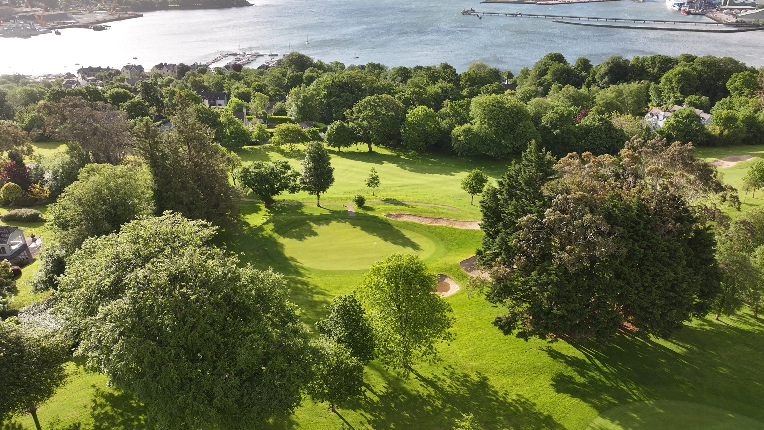 Aerial view of a golf course near a body of water with trees and residential houses nearby.