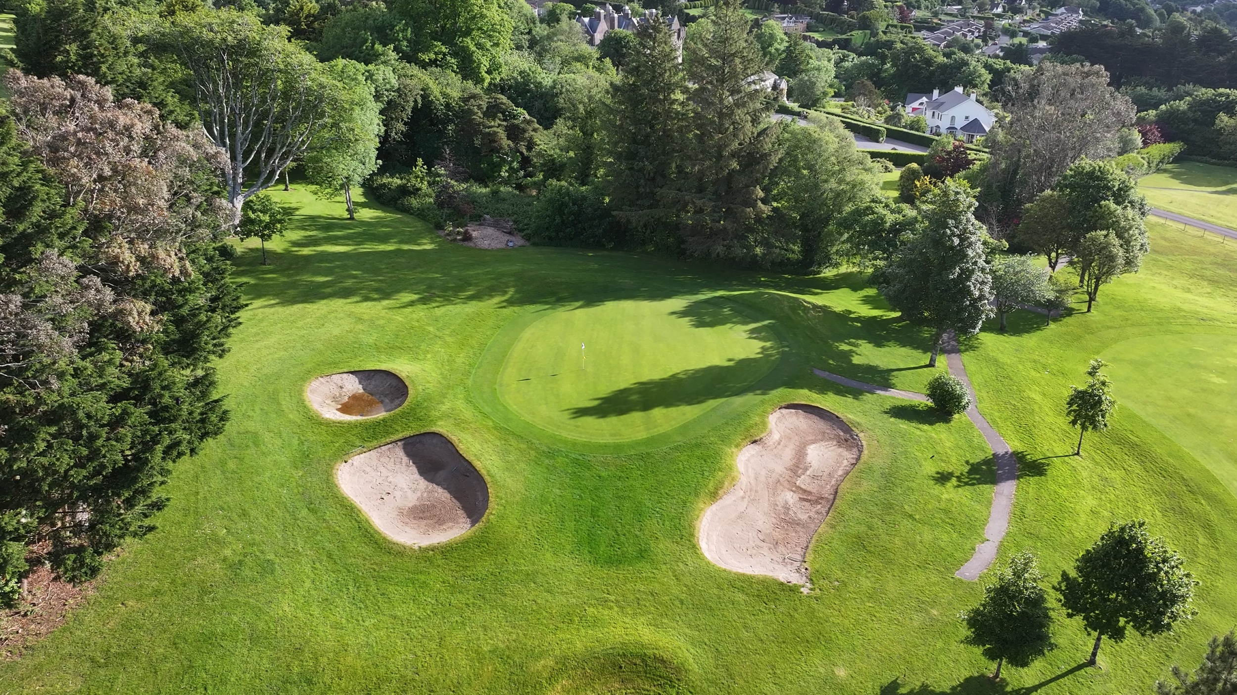 Aerial view of a golf course with a putting green, surrounded by sand bunkers, trees, and a pathway, with residential houses in the background.
