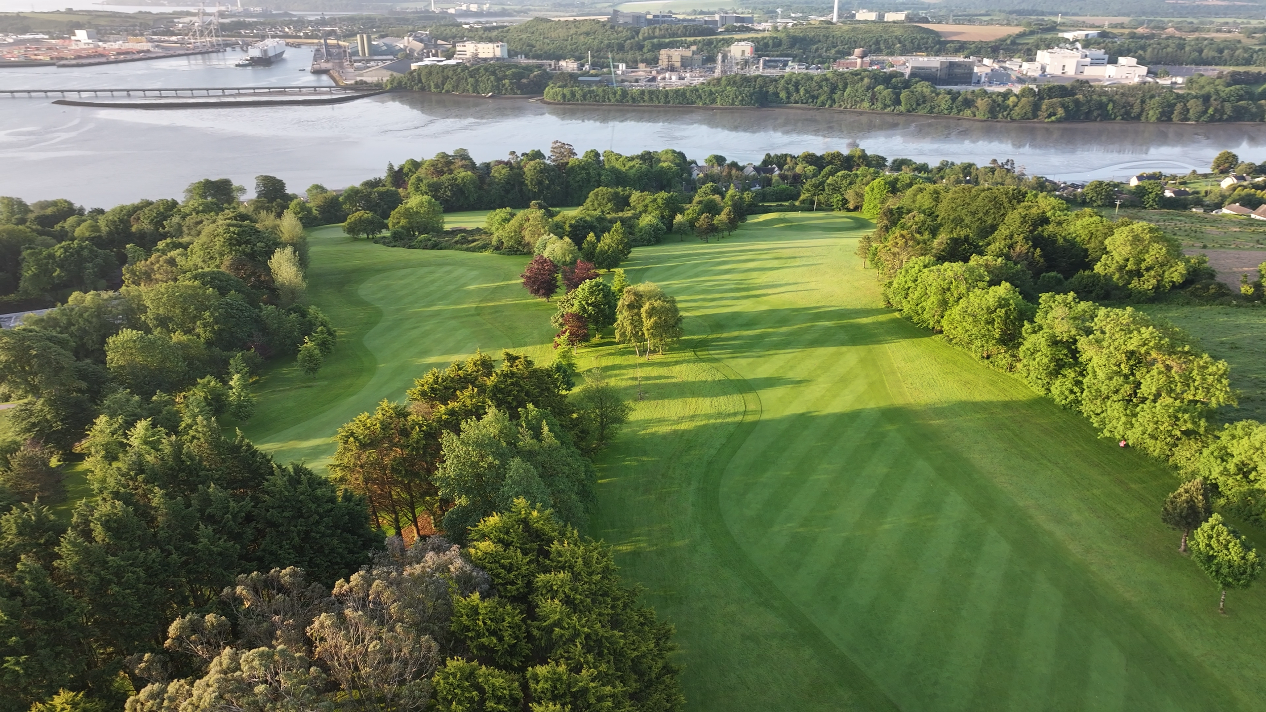 Aerial view of a golf course with neatly mowed green fairways and mature trees, adjacent to a river with industrial buildings and a bridge in the background under a clear sky.