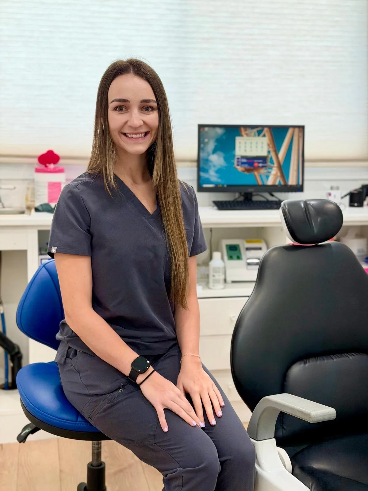 A smiling woman in gray scrubs sitting on a blue medical stool in a clinical office setting with medical equipment and a computer monitor in the background.