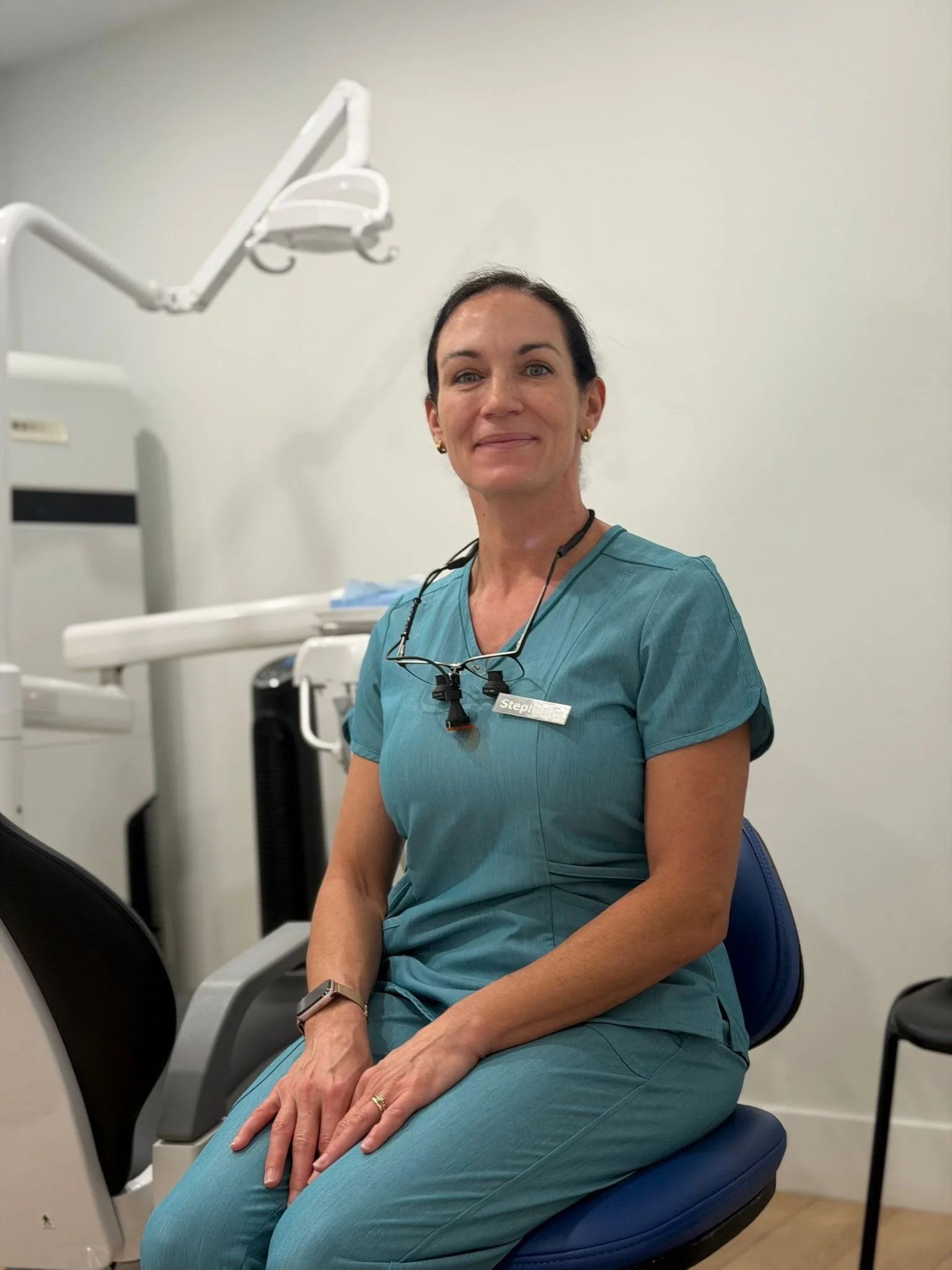 A woman in teal medical scrubs sitting in a doctor's office, smiling at the camera. She is wearing glasses hanging from her neck and a name tag that says 'Stephanie.' There is medical equipment in the background.
