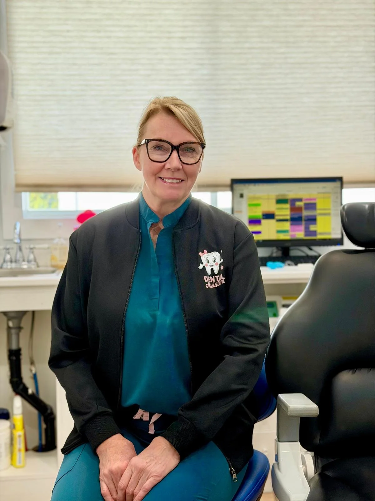A smiling woman with glasses and short blonde hair sitting in a dental office, wearing a black jacket with an embroidered tooth and the words 'Dental Assist' on it. Behind her is a computer monitor displaying a colorful schedule, and dental tools are visible in the background.