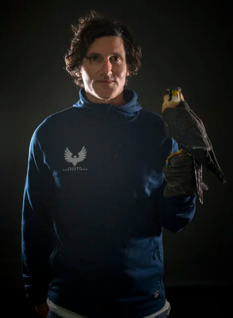 A man standing against a dark background, holding a bird of prey on his gloved hand.