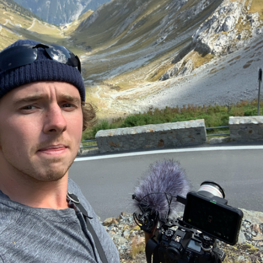 A man taking a selfie with a camera and microphone on a mountain road, surrounded by rocky slopes and green grass.