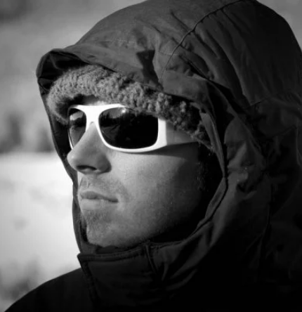 Profile of a man wearing a hooded jacket, white sunglasses, and a beanie outdoors in winter