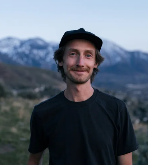 A man with a beard and mustache wearing a black t-shirt and a black cap, standing outdoors with snow-capped mountains in the background on a clear day.