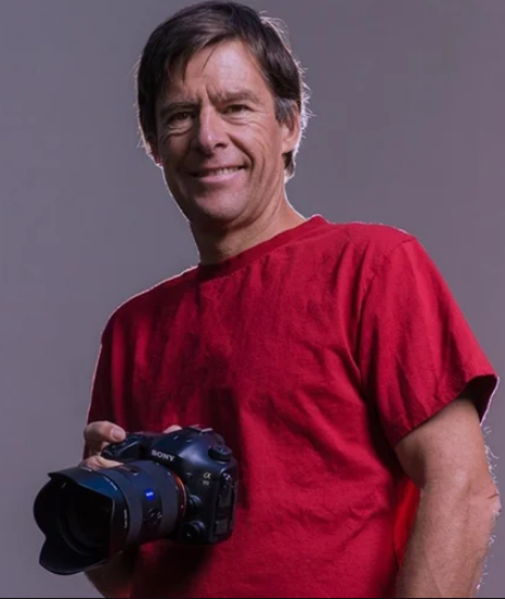 A man smiling while holding a professional camera, wearing a red t-shirt against a plain gray background.