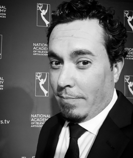 Black and white portrait of a man with curly dark hair, wearing a tuxedo and tie, standing in front of a backdrop with the logo of the National Academy of Television Arts & Sciences.