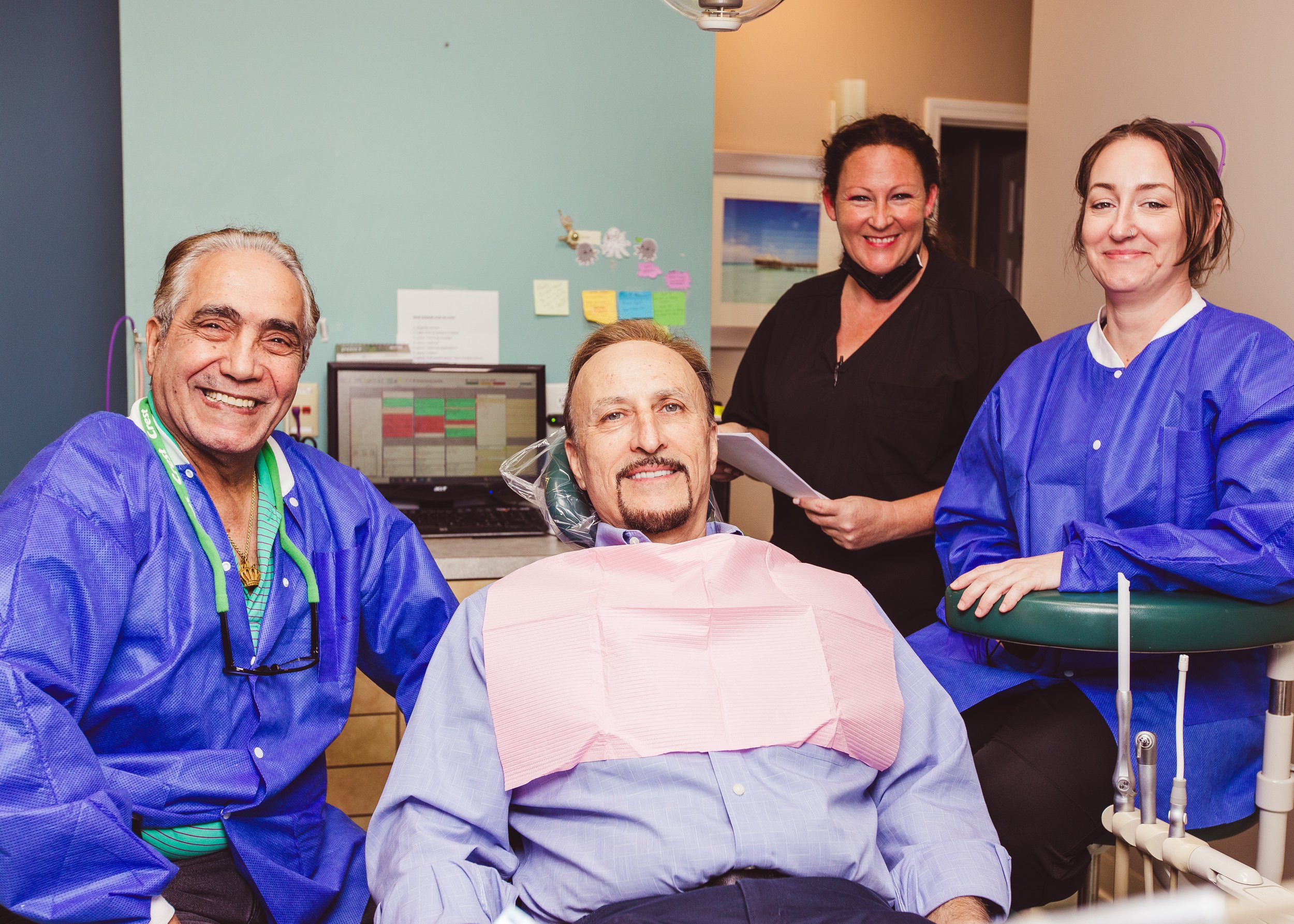 A smiling older man in a blue medical gown sitting in a dental or medical office with a dental chair and equipment, indicating he is a patient.