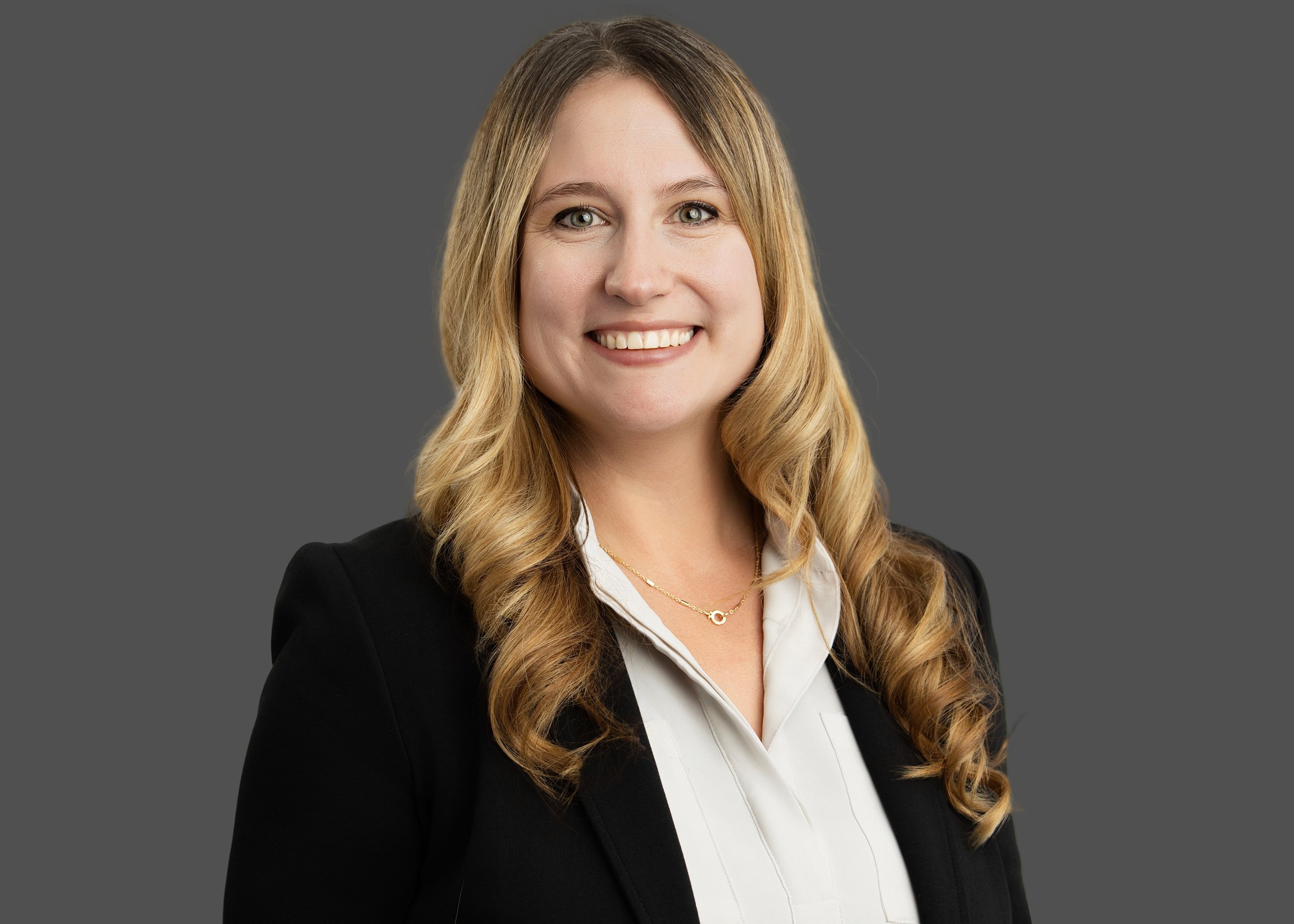 Professional woman with long, wavy hair, smiling, wearing a black blazer and white blouse, against a gray background.