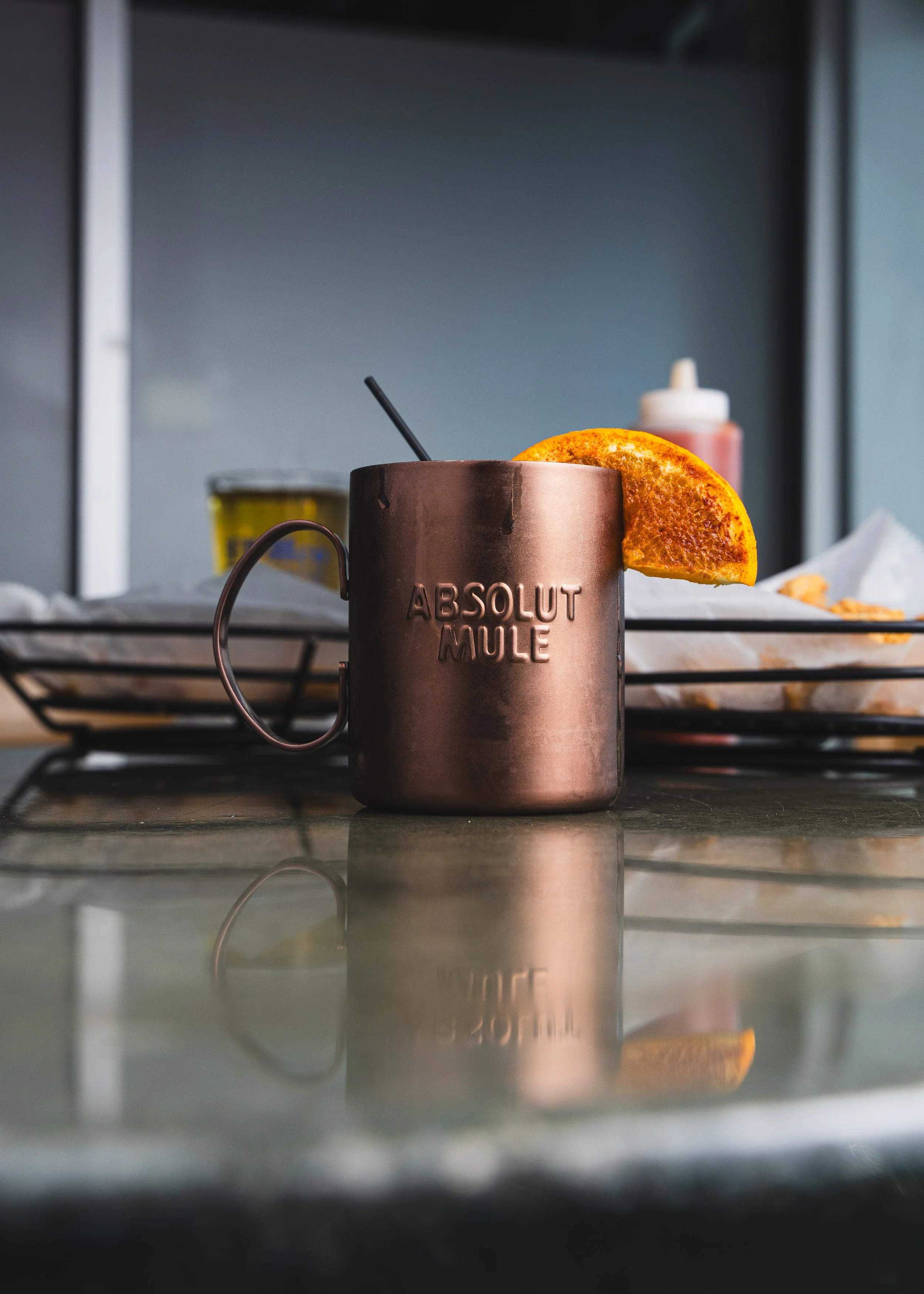 Close-up of a copper mug with the words "ABSOLUT MULE" embossed on it, garnished with an orange slice. In the background, a bottle of hot sauce, mustard, and a paper towel on a wire rack are visible.