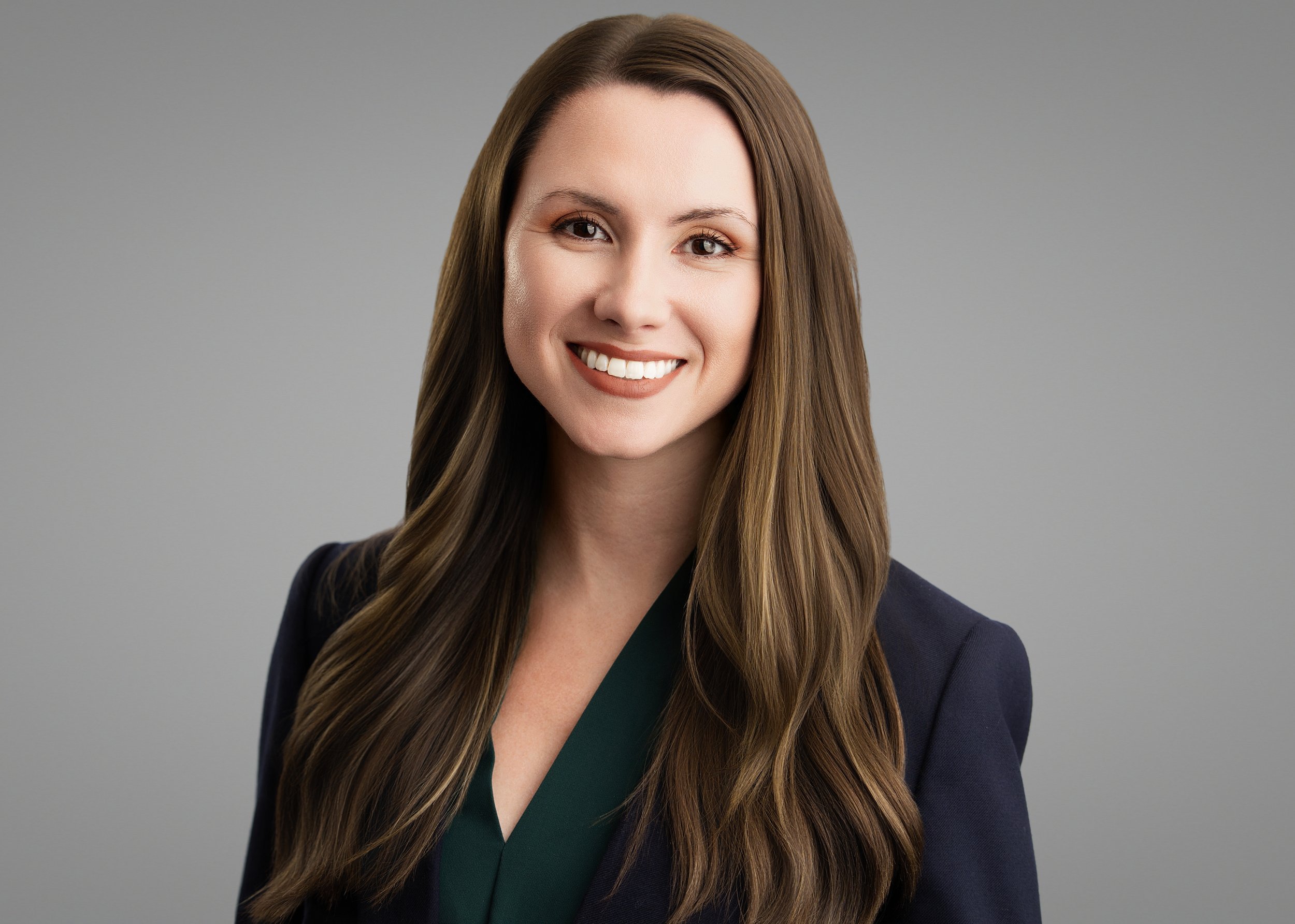 A woman with long brown hair styled in loose waves, wearing a dark blazer and a green top, smiling against a plain gray background.