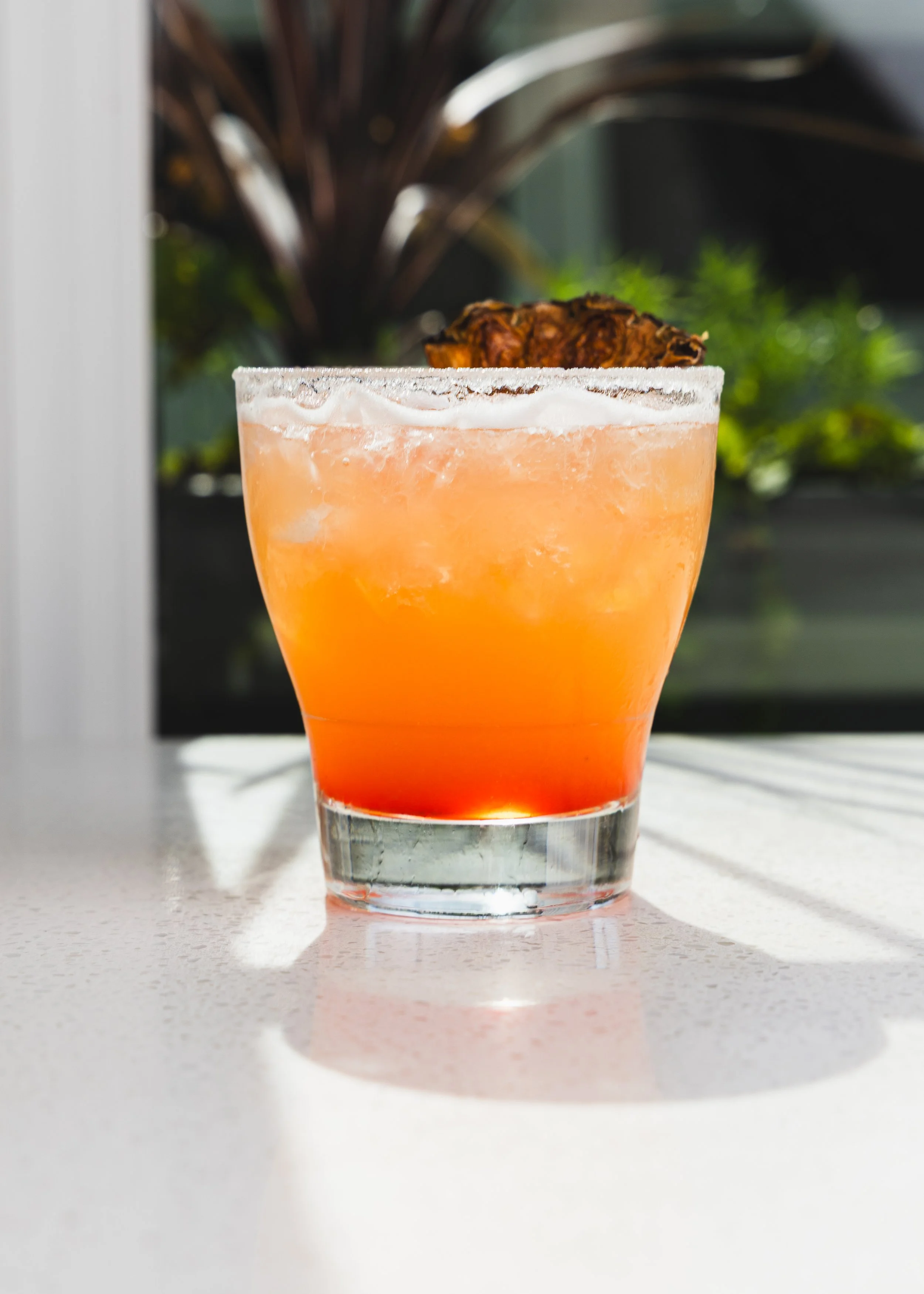 Close-up of an orange cocktail in a clear glass on a white surface with blurred plants in the background.