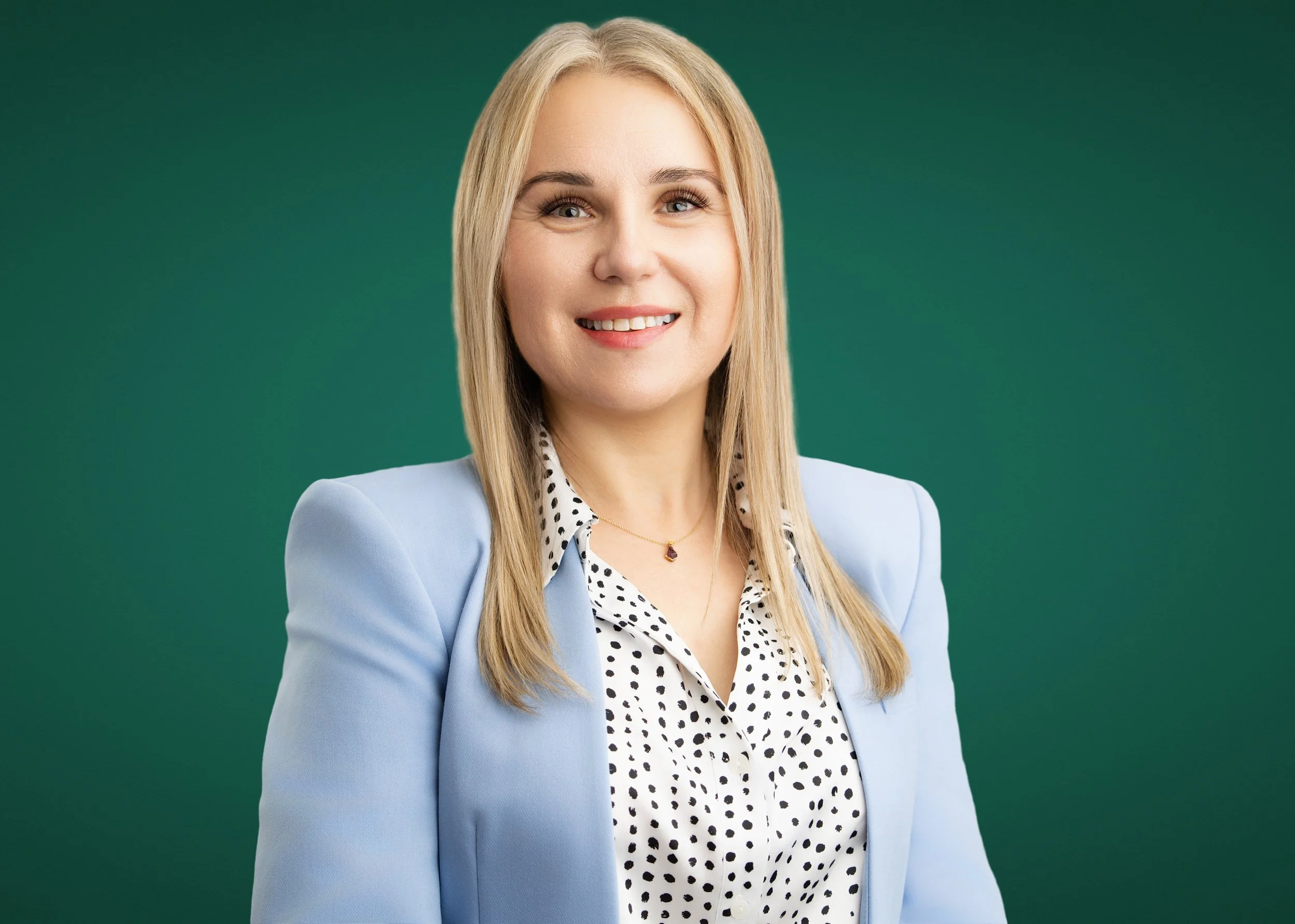 A smiling blonde woman wearing a light blue blazer and a black and white polka dot blouse; standing against a green background.