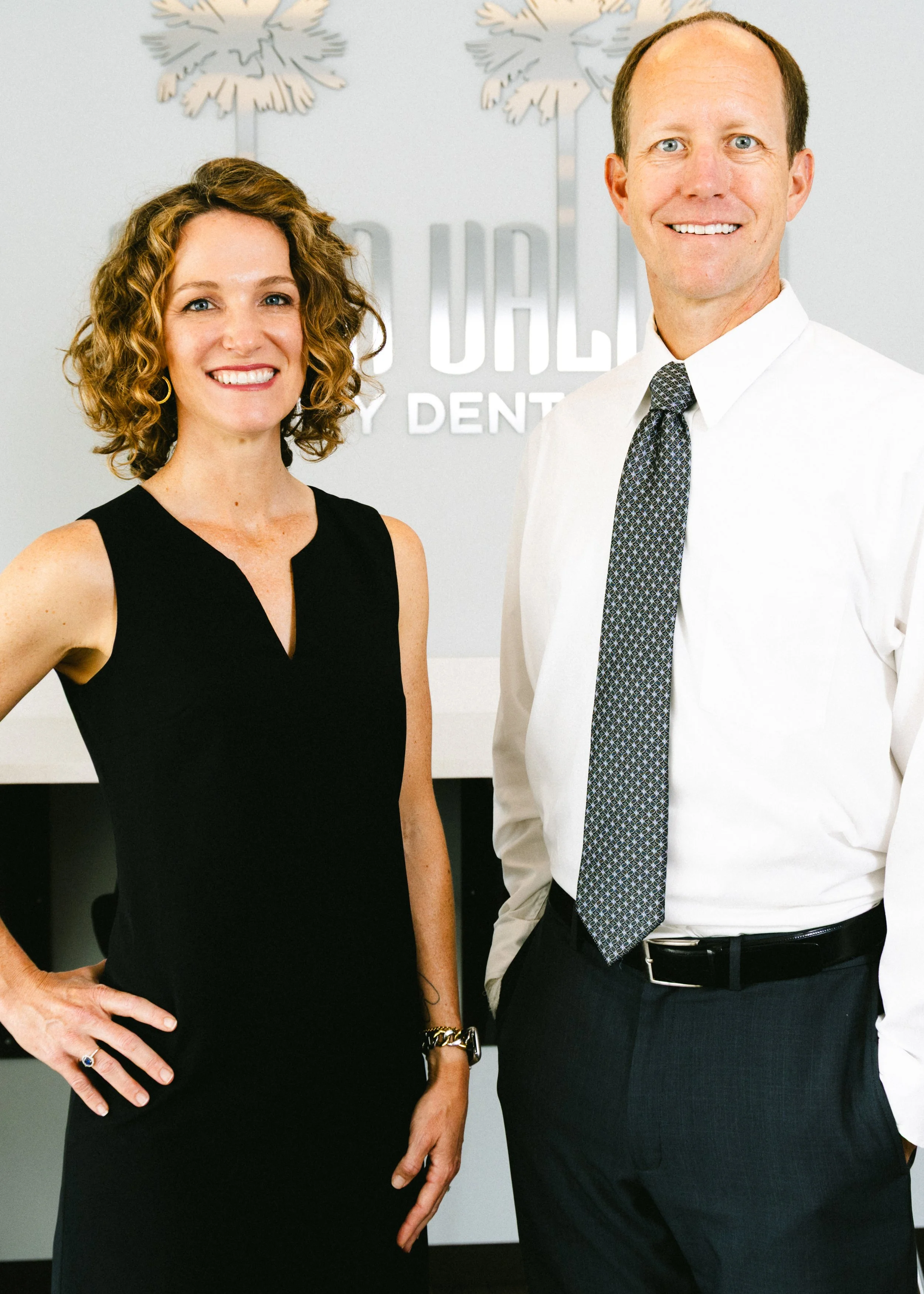 Two professionals, a woman with curly brown hair and a man with short brown hair, standing in front of a logo that reads 'Doherty Family Dentistry', smiling at the camera.