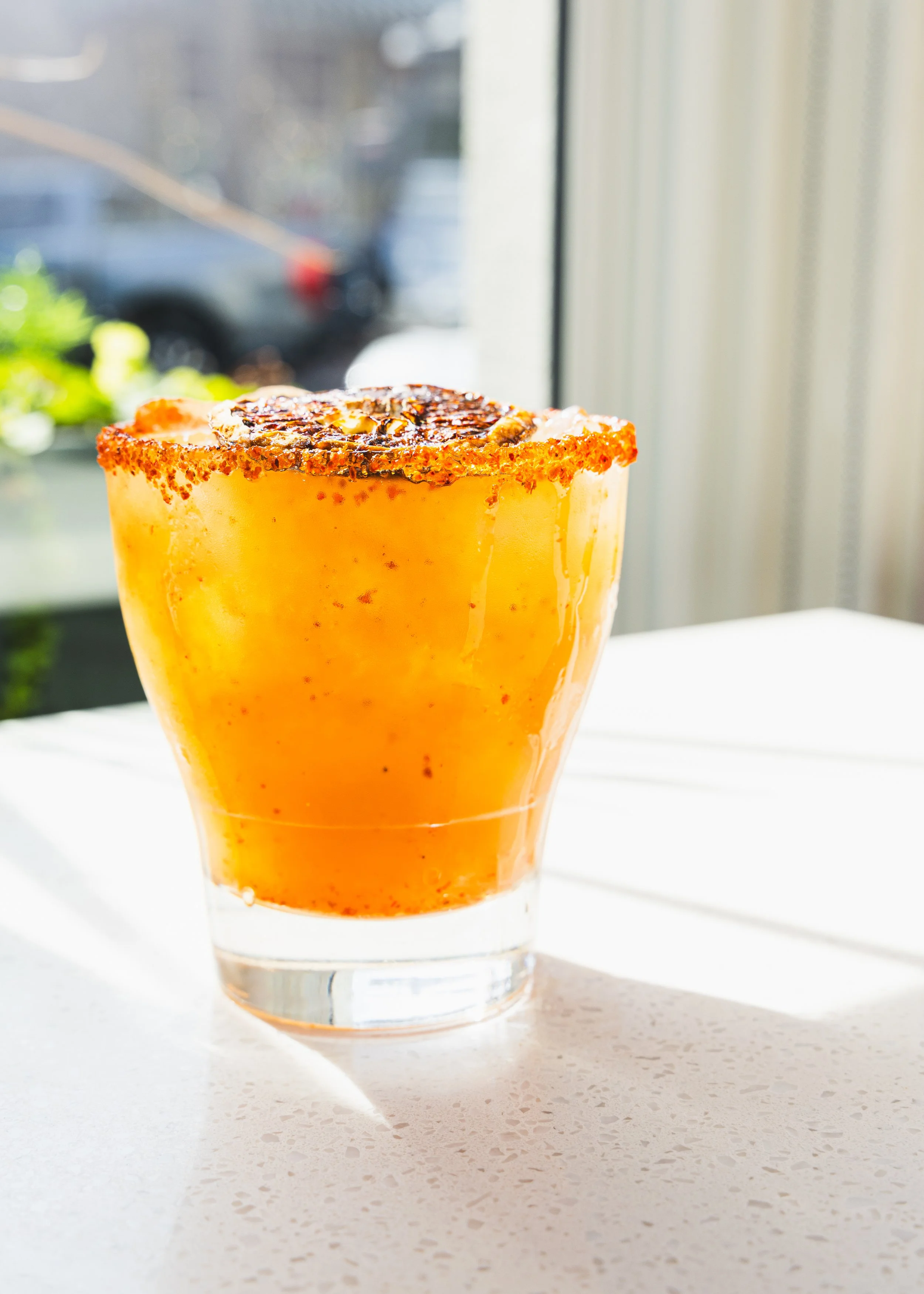 A close-up of a glass containing a yellow-orange beverage with a spicy rim and a charred lemon slice on top, sitting on a white surface near a window with sunlight.