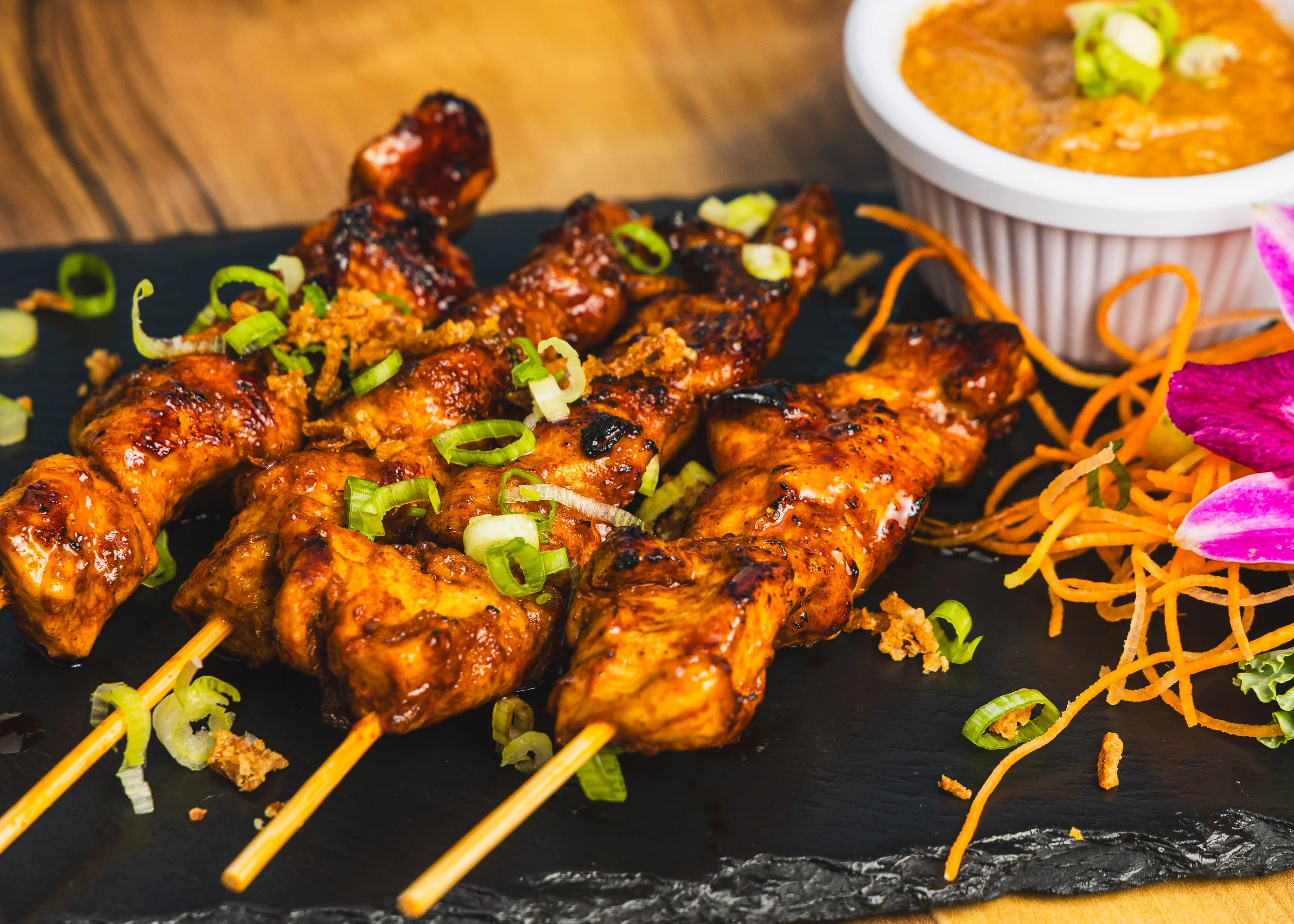 Close-up of grilled skewers of chicken garnished with chopped green onions, served on a black plate with colorful side dishes and a bowl of curry in the background.