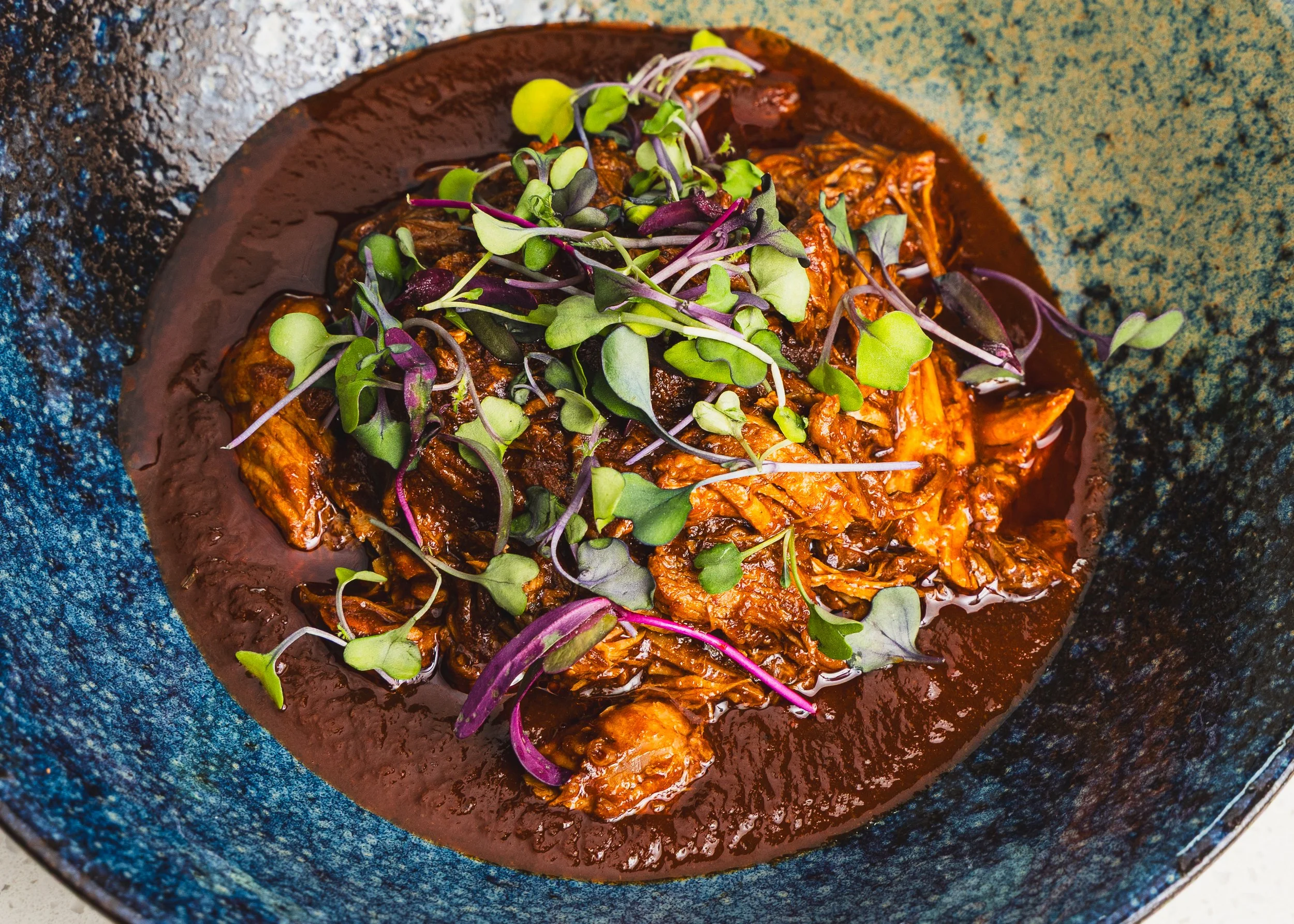 A dark brown dish of shredded meat topped with microgreens, served in a black bowl with a textured blue interior.