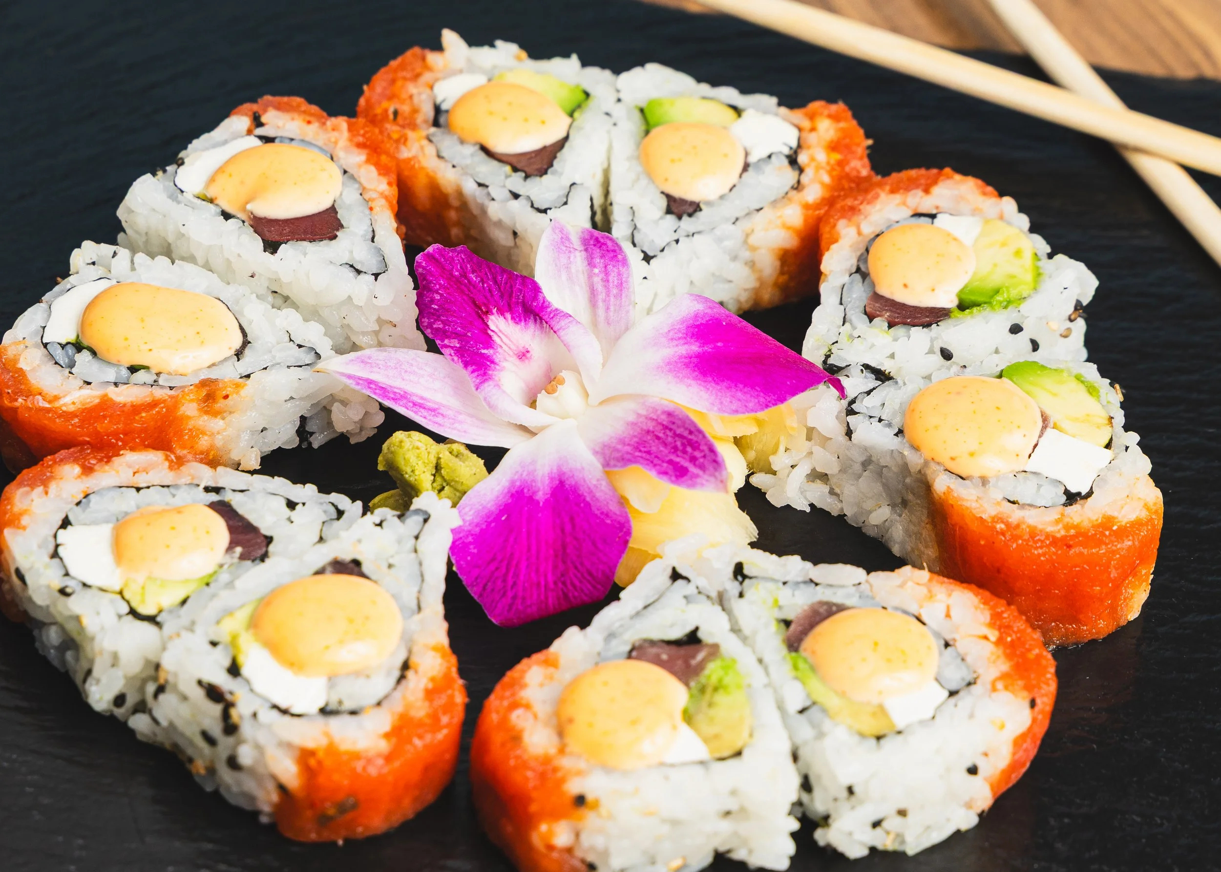 Sushi rolls with fish, avocado, and egg, garnished with a pink orchid flower on a black surface.