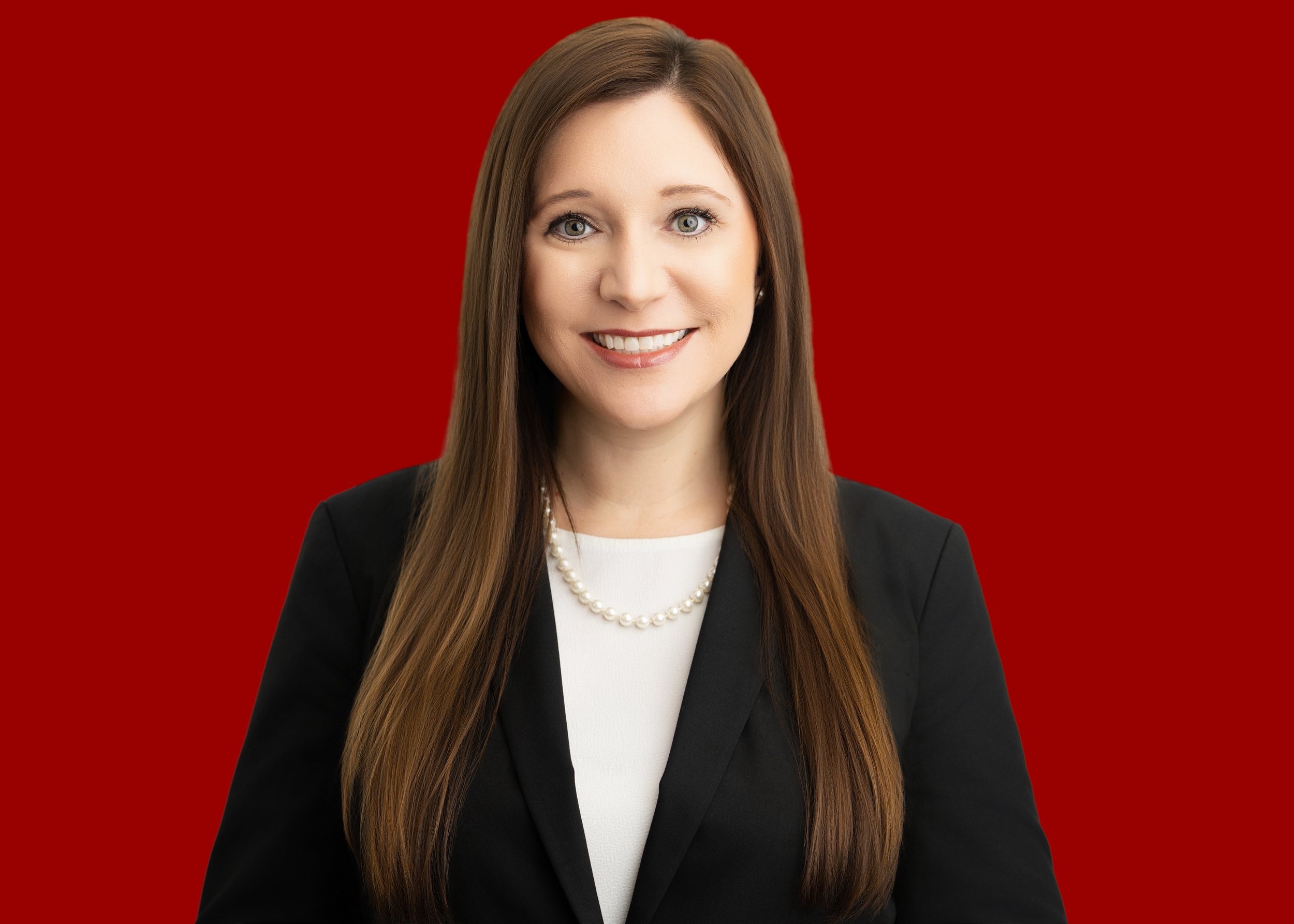 A professional woman with long brown hair, wearing a black blazer, white top, pearl necklace, and earrings, smiling against a solid red background.