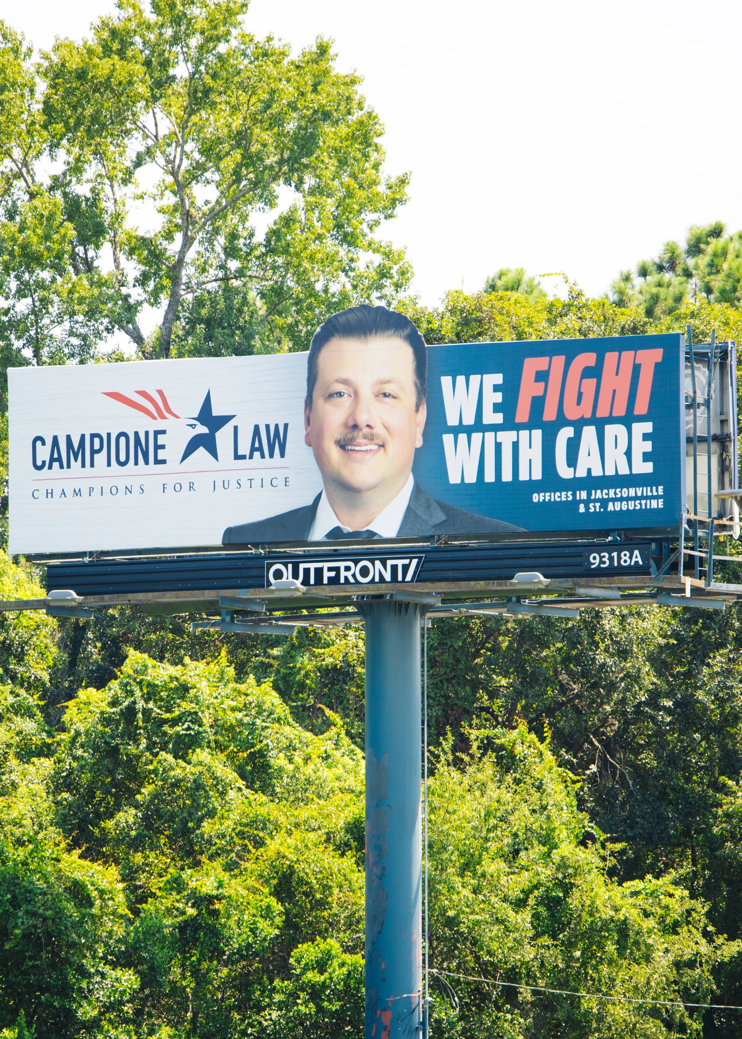 A large billboard with a photograph of a man with dark hair, smiling, wearing a suit. The billboard displays the text 'Campione Law', 'Champions for Justice', and 'We Fight With Care'. It also mentions offices in Jacksonville and St. Augustine.