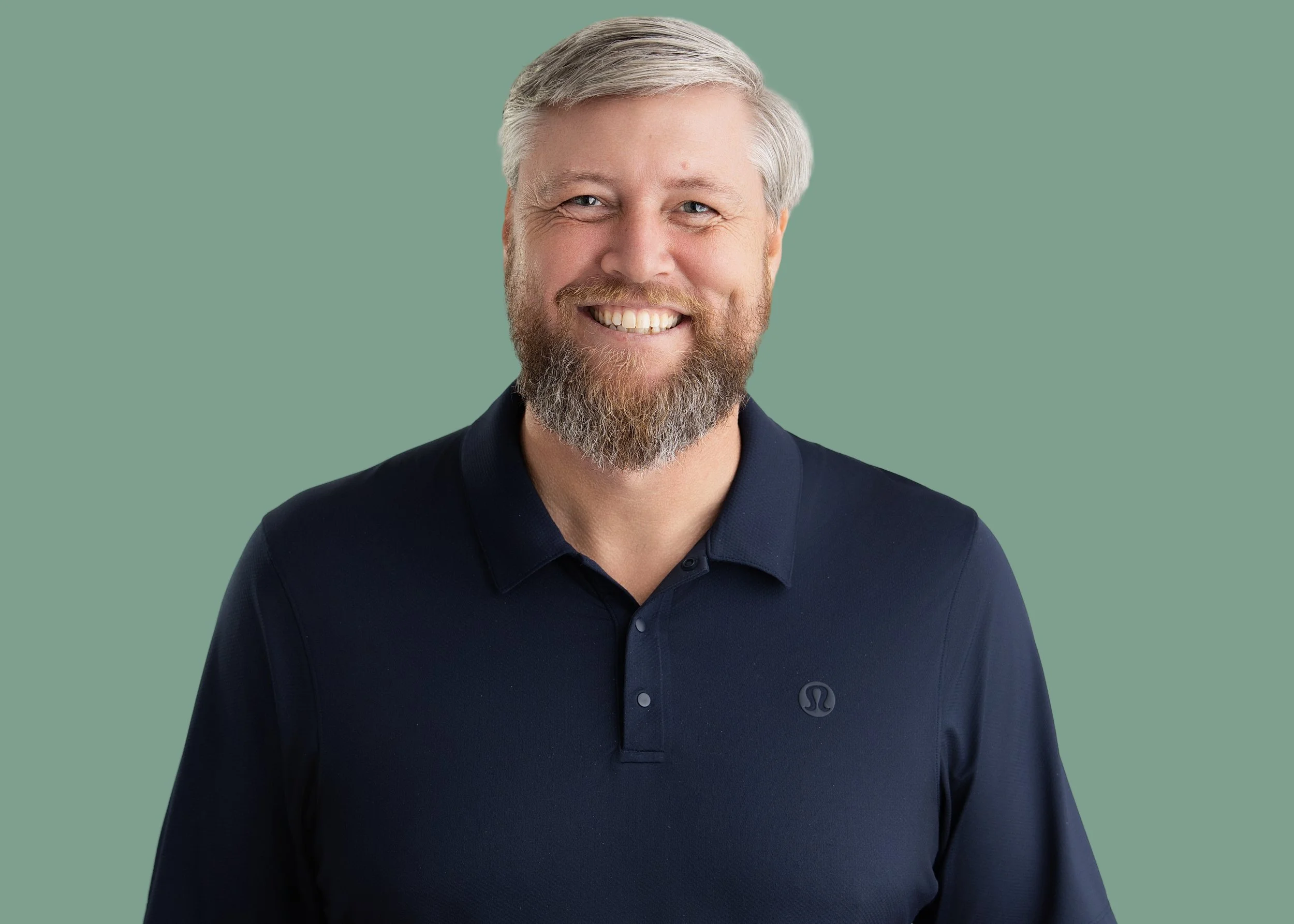 Photo of a smiling middle-aged man with gray hair and a beard wearing a dark polo shirt, standing against a muted green background.