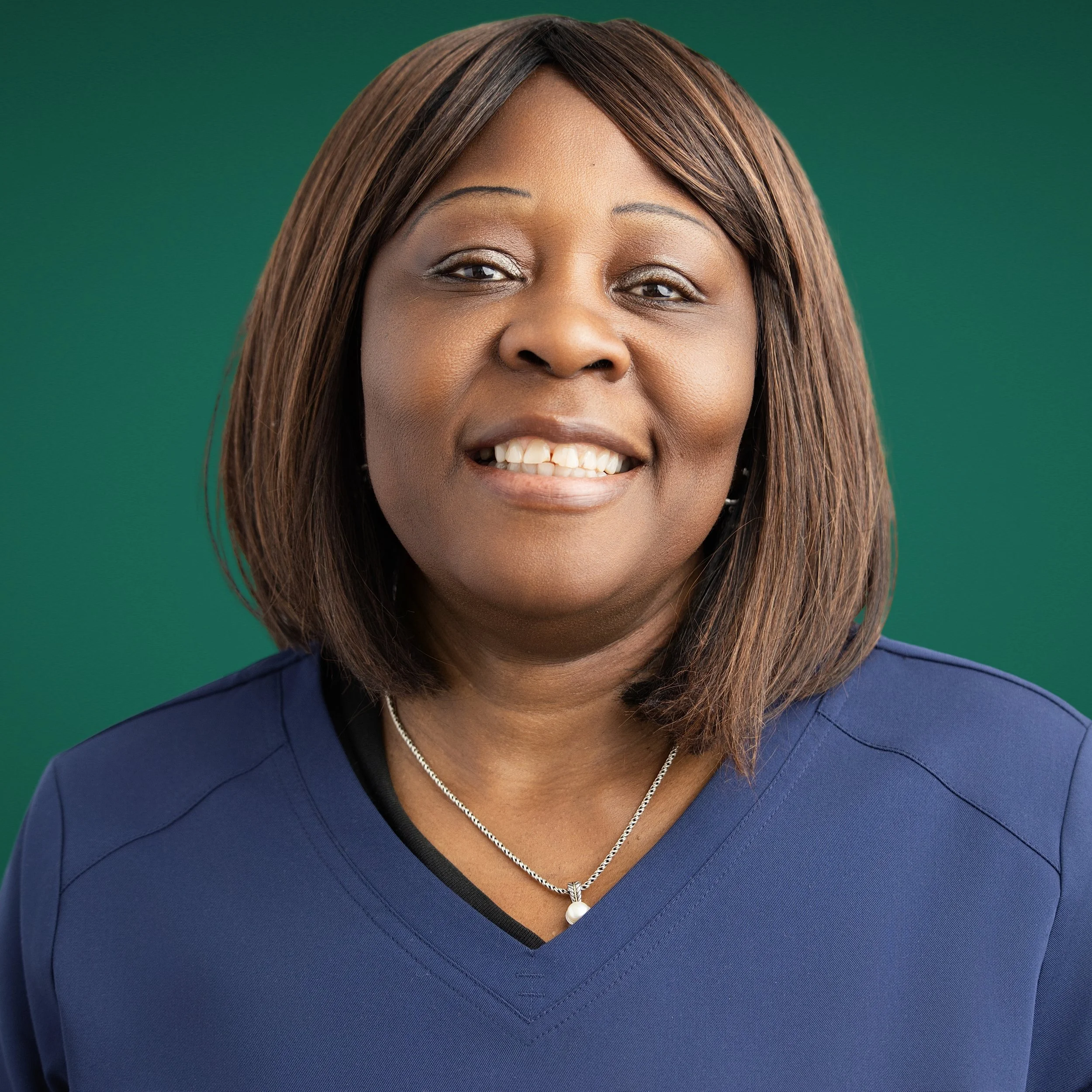 Close-up of an African American woman's face smiling, with brown hair, wearing a blue top, a pearl necklace with a knot and pearl pendant, and with a green background.