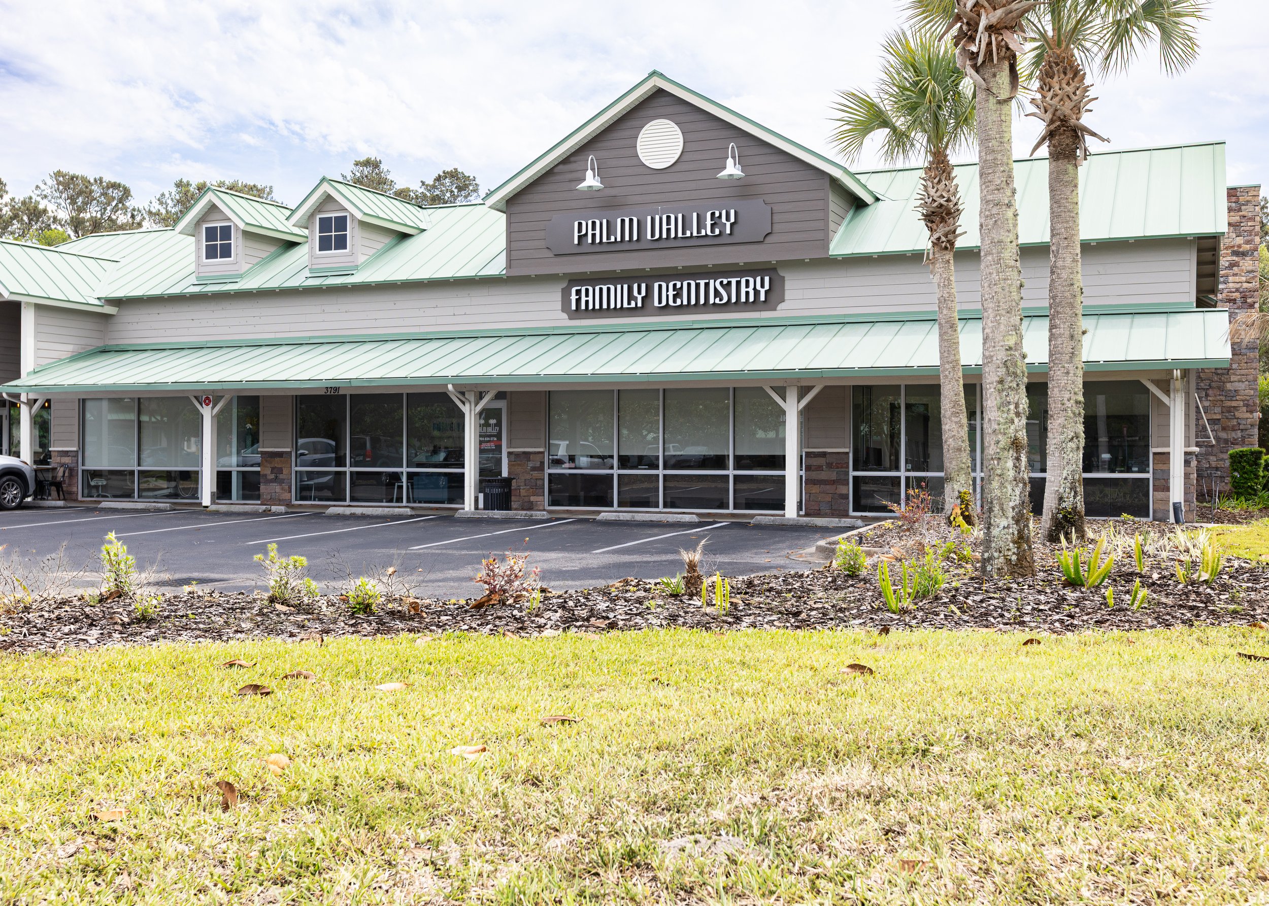 Exterior of a building with a sign that reads 'Palm Valley Family Dentistry,' with a green metal roof, large windows, and three tall palm trees in front, under a partly cloudy sky.