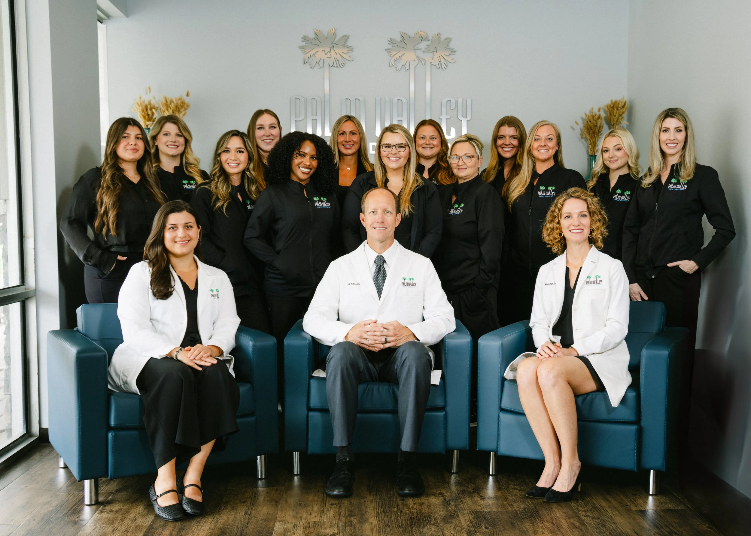 Group photo of fifteen people, mostly women, in a modern office setting. They are smiling, some standing and some sitting, with a light gray wall behind them displaying the text "PALM VALLEY" and a palm tree logo, and a large window to the left. Four women are seated in front, wearing white coats, while the others are standing in black shirts with the logo.