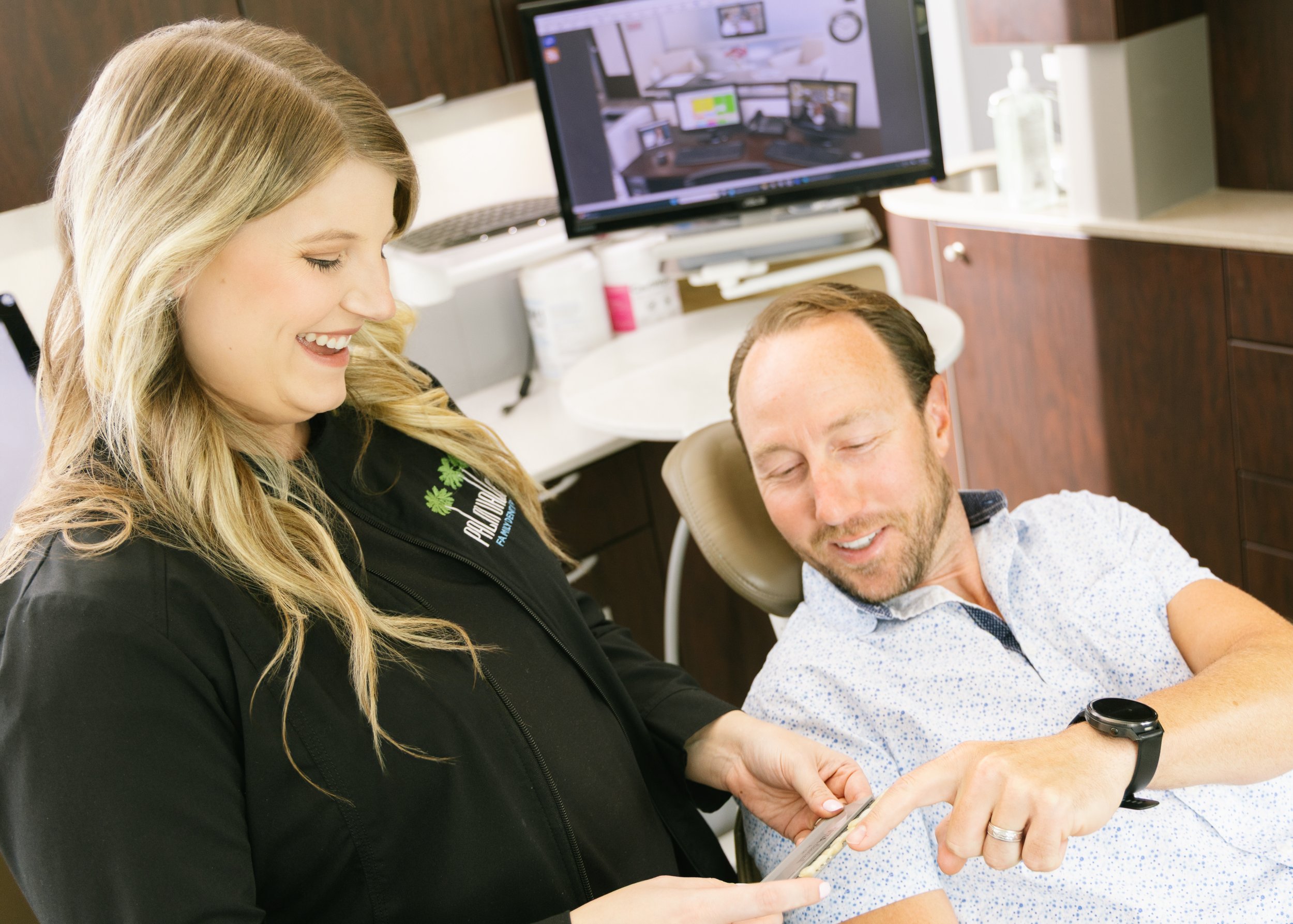 A woman smiling at a man who is lying in a dental chair, both looking at a smartphone.