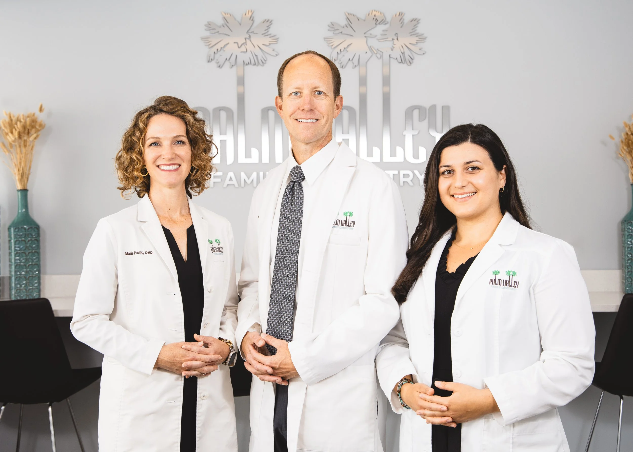 Three medical professionals in white coats standing in an office, smiling at the camera, with a sign and decorative vases visible in the background.