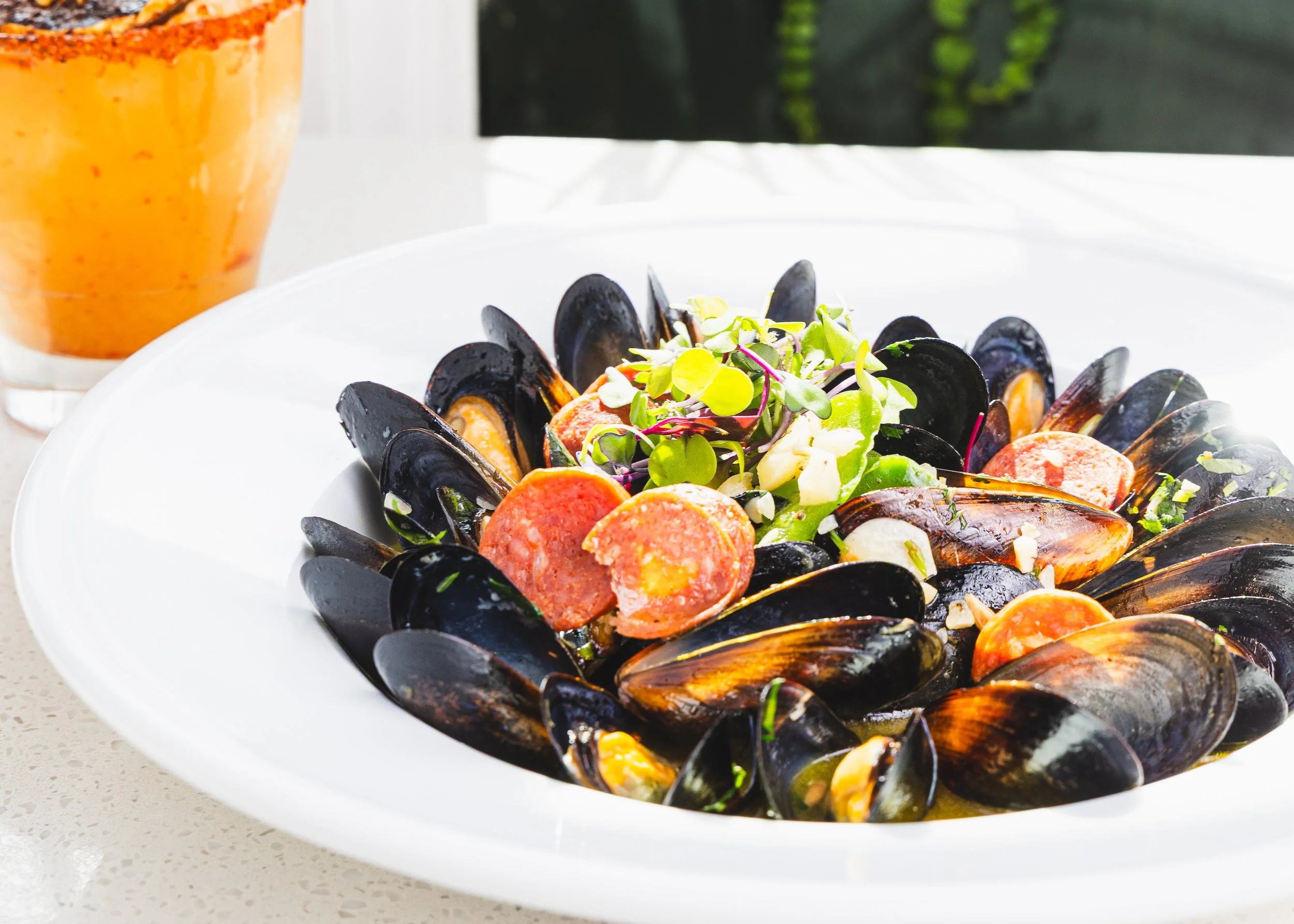A white plate with cooked mussels and slices of sausage, garnished with microgreens and minced garlic, with a glass of orange-colored beverage behind it.