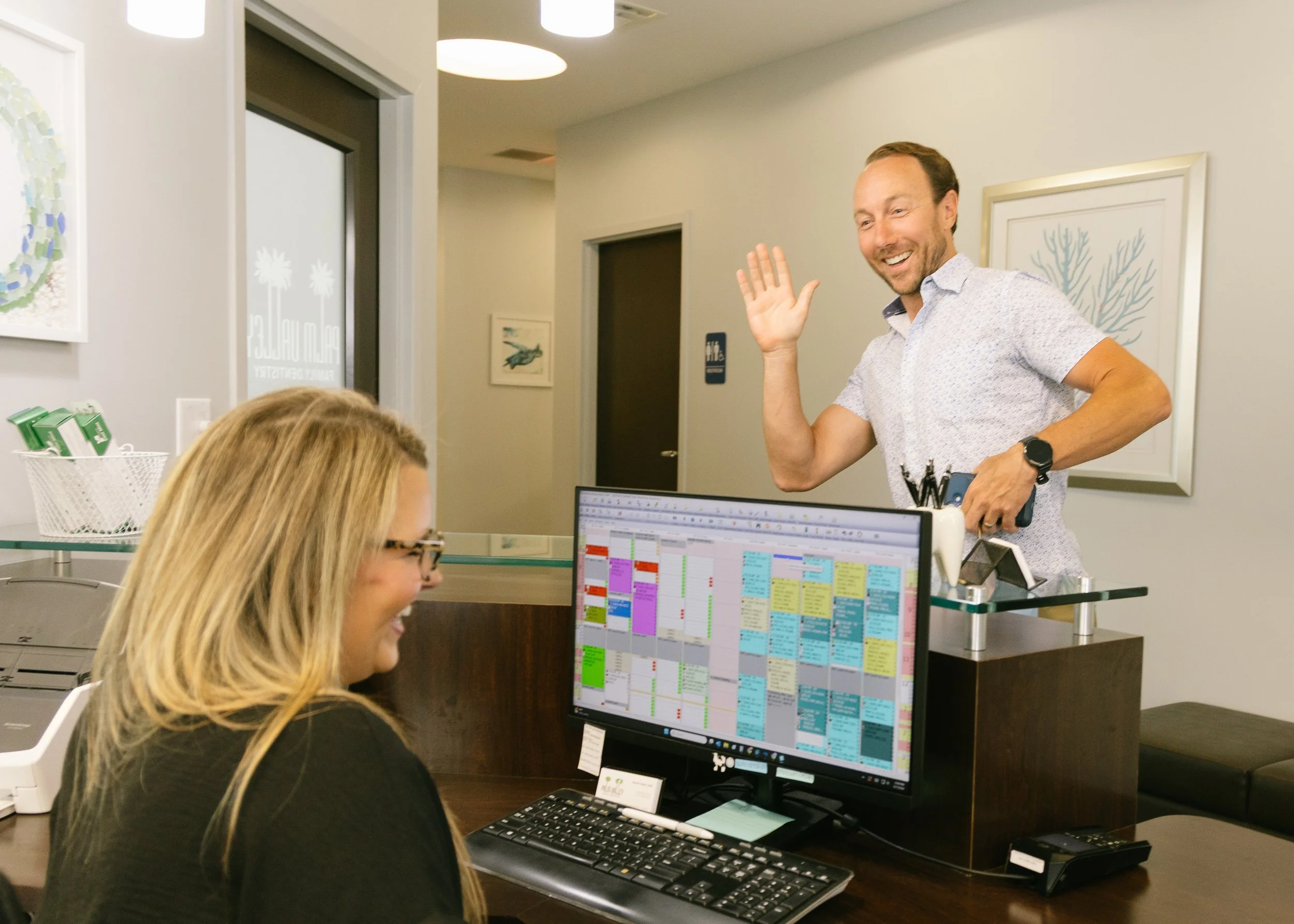 A man with a beard and light-colored hair smiling and waving while standing in an office room against a wall with framed artwork. He is wearing a short-sleeved, patterned shirt and a watch on his left wrist. In the foreground, a woman with blonde hair and glasses is working on a computer with a colorful schedule on the screen. The room has light-colored walls, ceiling lights, and decorated with artwork, including sea creatures and abstract designs.