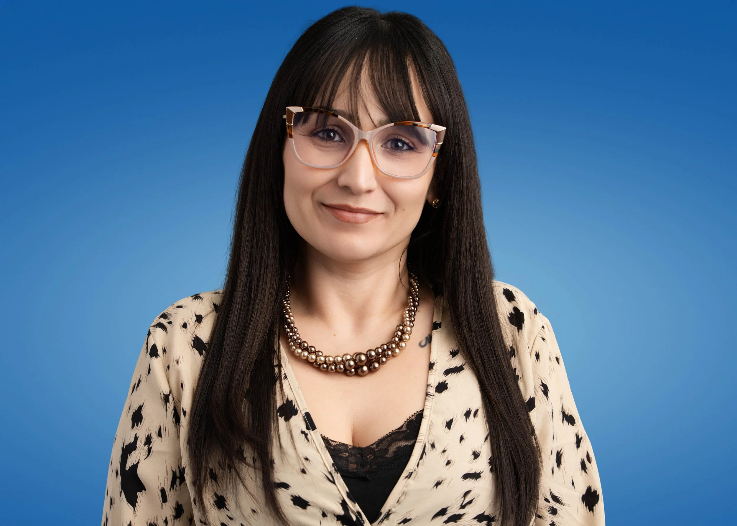 A woman with long dark hair, wearing glasses with a tortoise shell pattern, a pearl necklace, and a beige blouse with black print, smiling against a solid blue background.