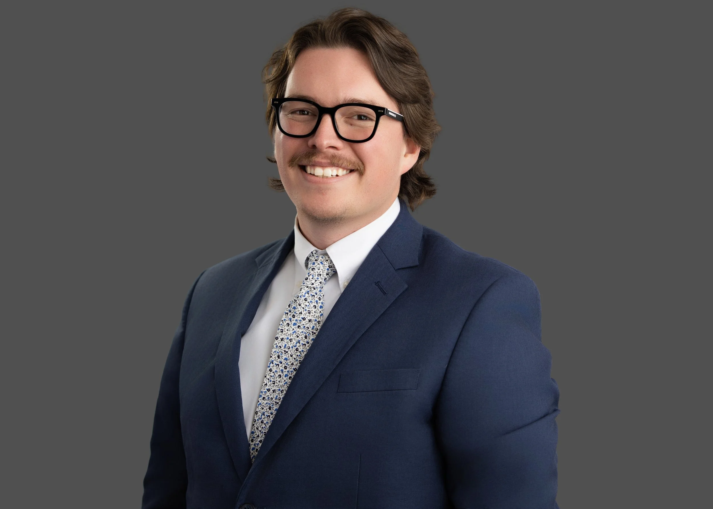 A smiling man in a navy suit, white shirt, patterned tie, and black glasses posing against a plain gray background.