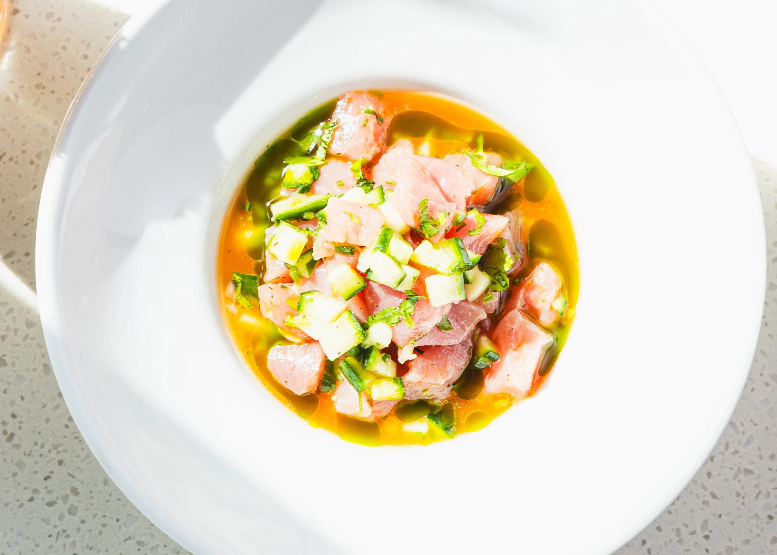A bowl of raw fish, possibly salmon, garnished with diced cucumbers and chopped herbs, topped with a drizzle of oil or sauce.