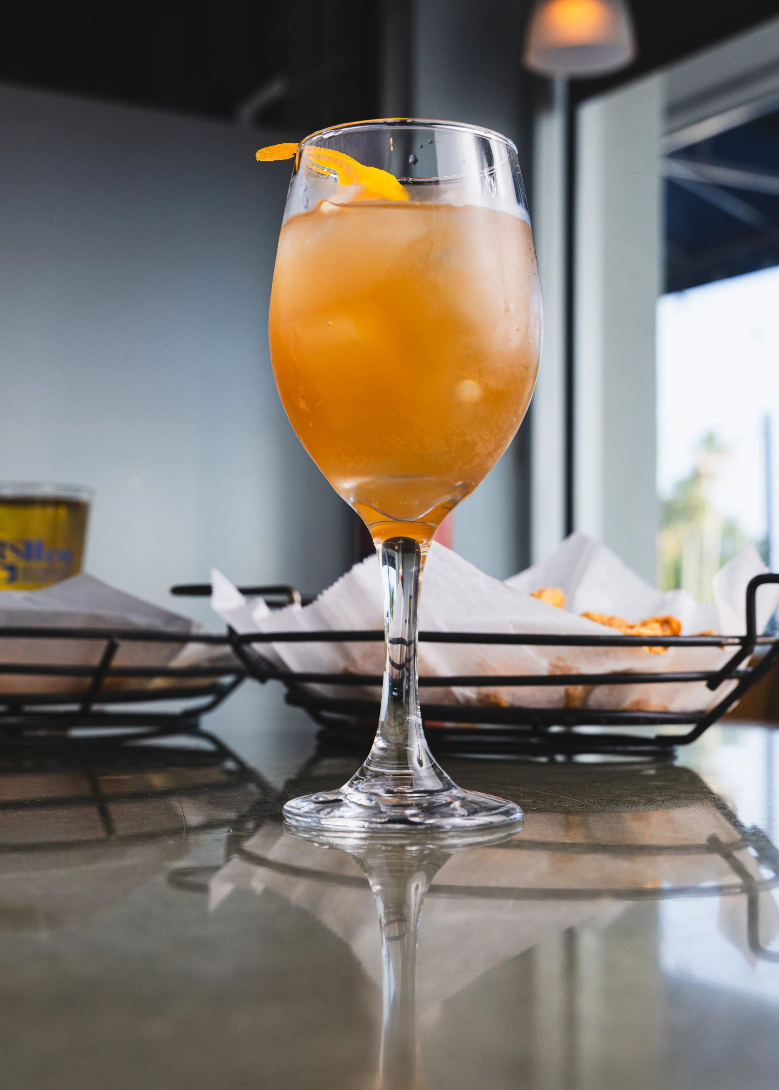 A cocktail glass filled with an orange-colored drink, garnished with a twist of orange peel, sitting on a reflective surface near a window with a dark interior background.