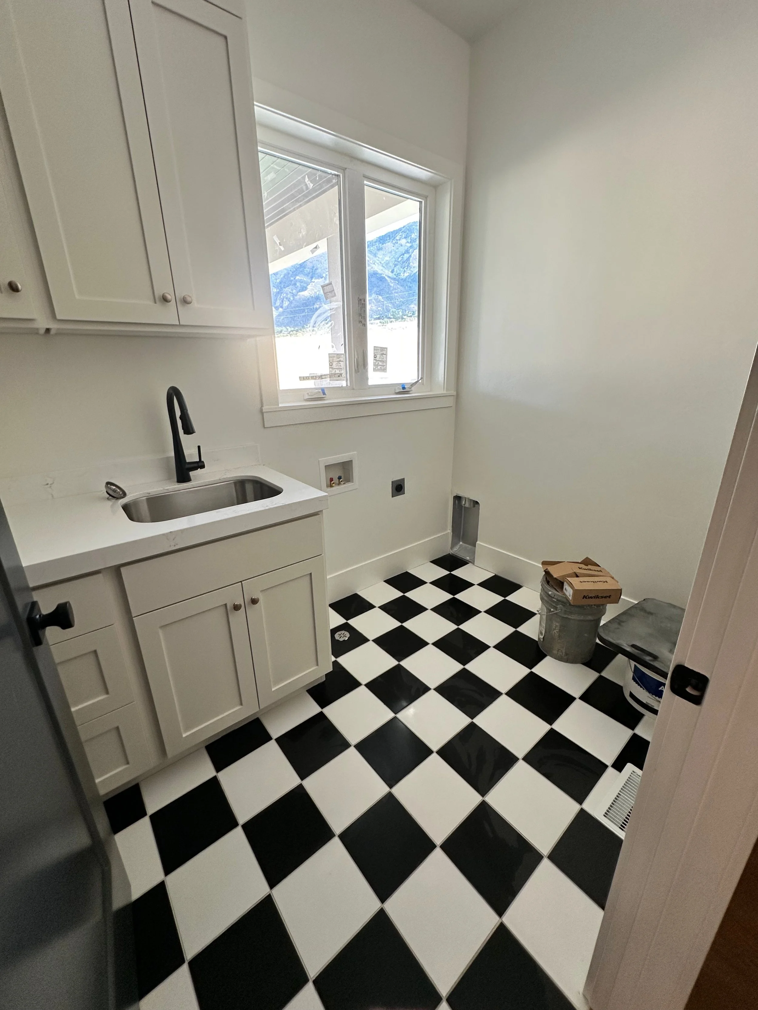Empty laundry room with black and white checkered floor, white cabinets, black sink faucet, window with mountain view, and construction supplies in corner.