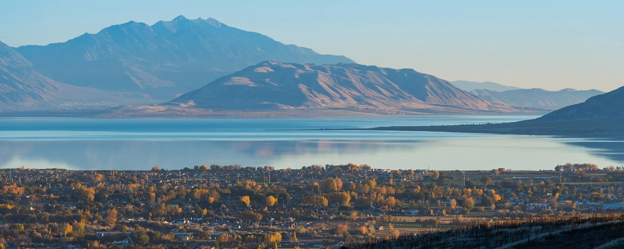 Landscape of a large lake with reflections, surrounded by trees with fall foliage, and distant mountains in the background under a clear sky.
