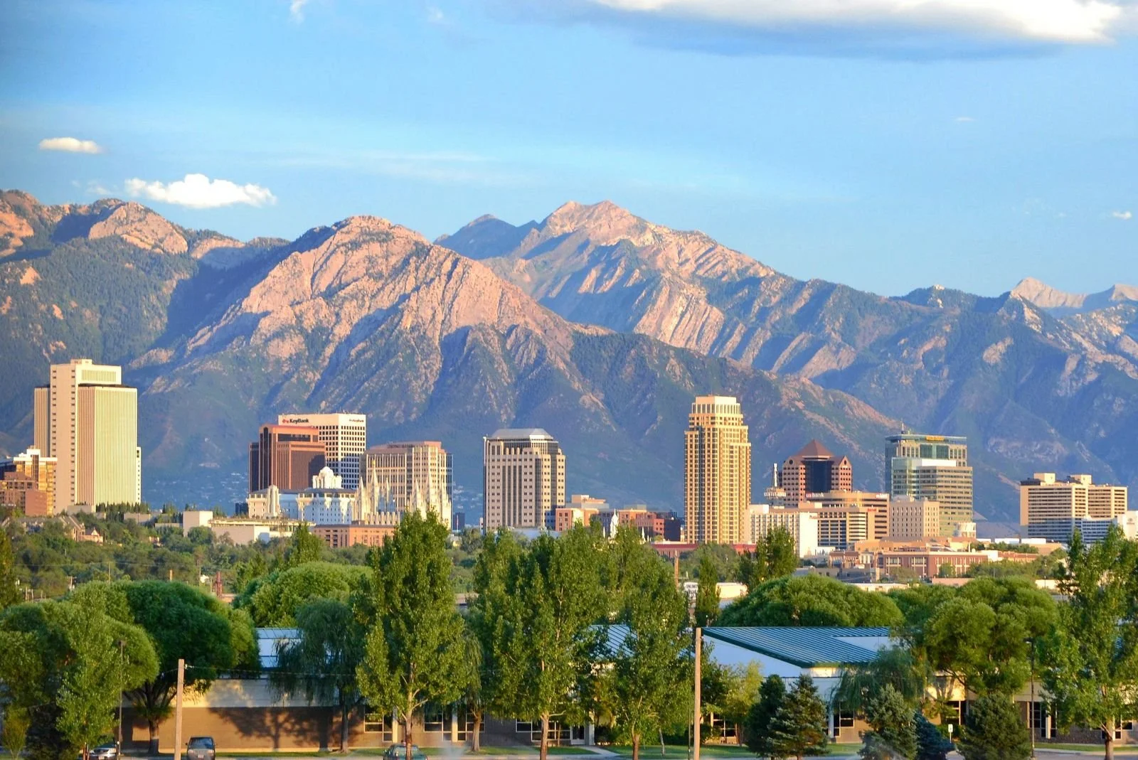 Cityscape with tall buildings in front of rugged mountain range, green trees, and partly cloudy sky.