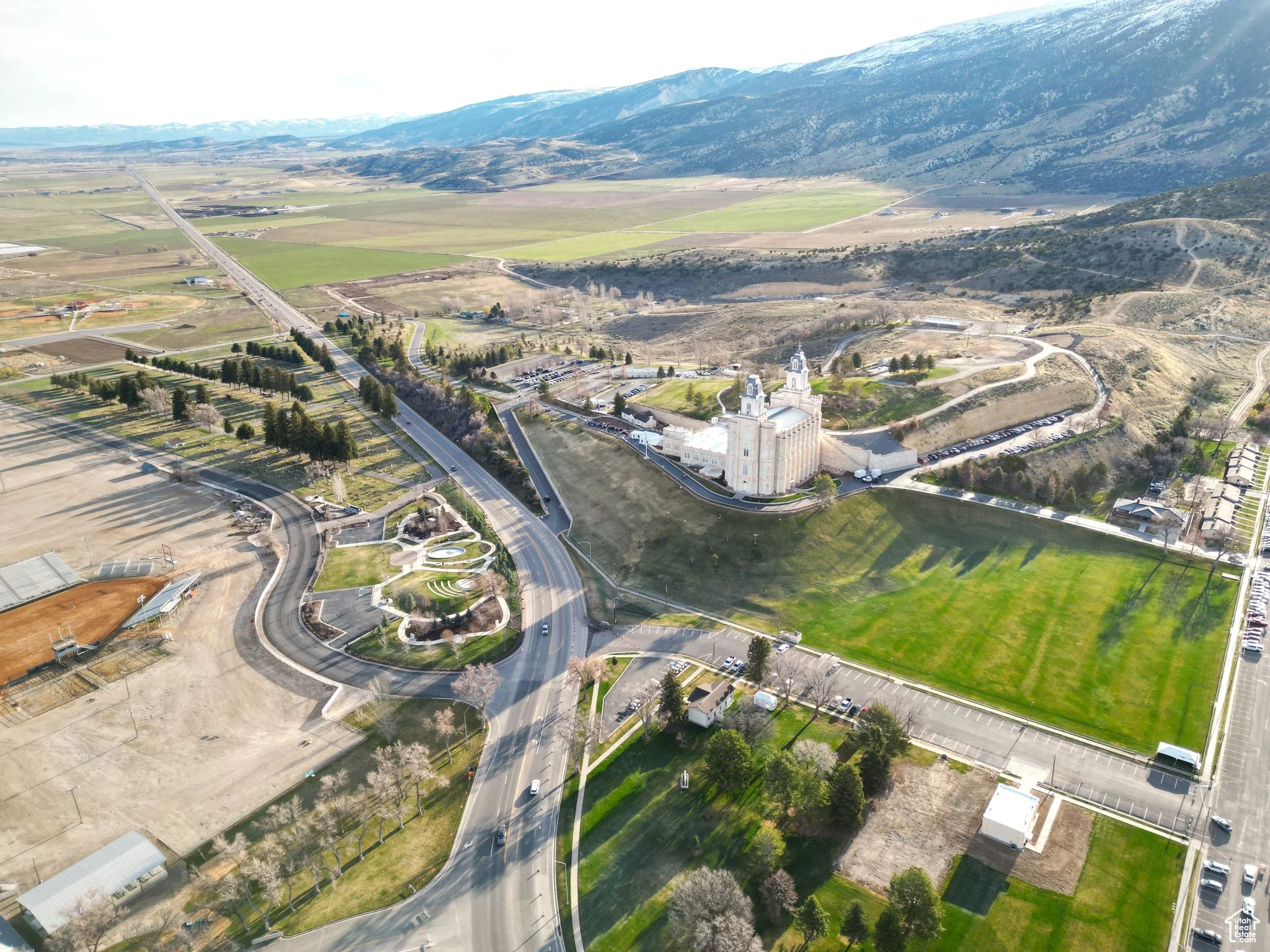 Aerial view of a historic church with twin towers, surrounded by parking lots, green grass fields, and mountainous terrain in the background.