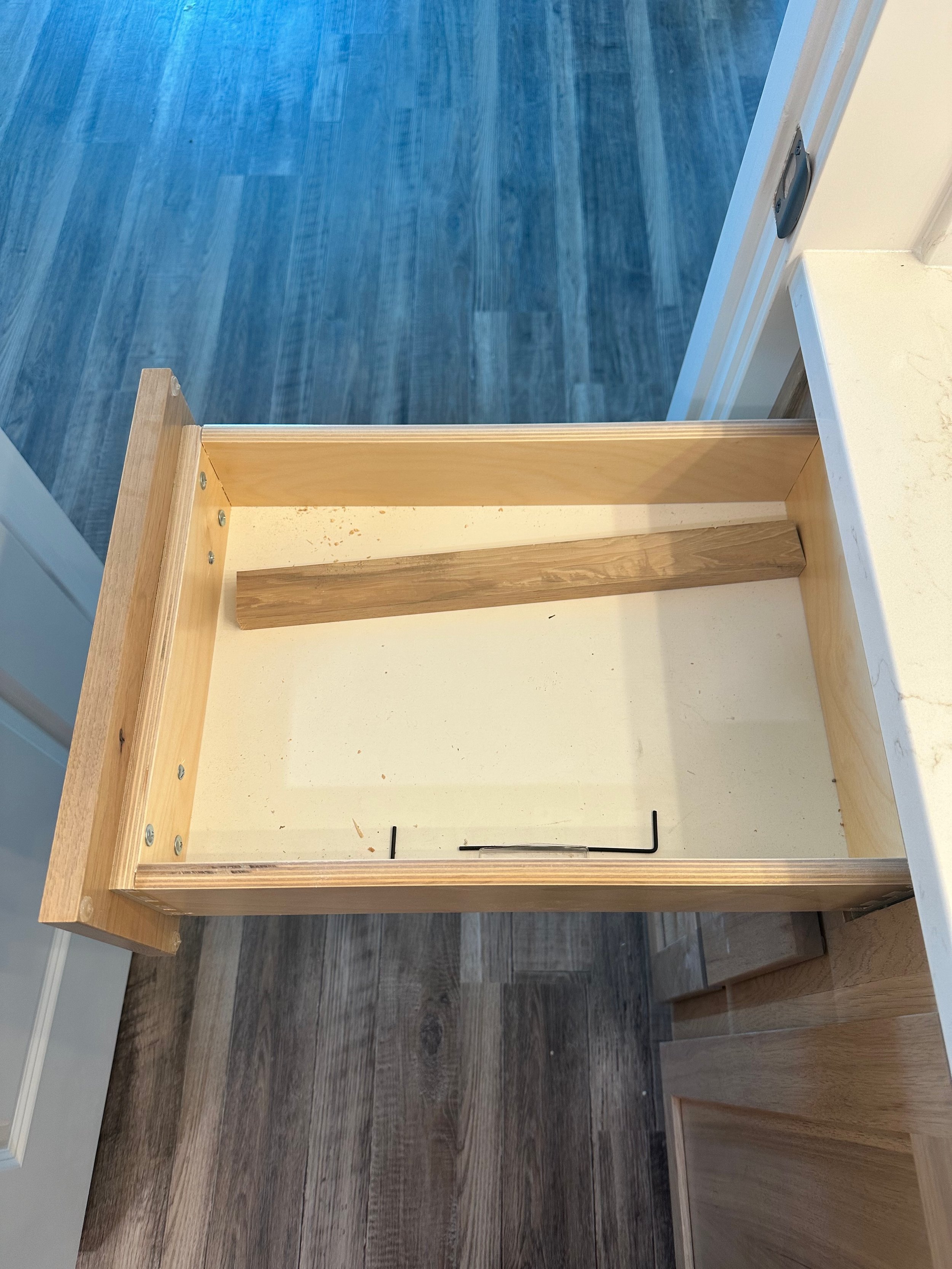 Unfinished wooden drawer with a metal handle, empty, inside a white cabinet or desk with partially visible white surfaces and a blue-colored flooring.