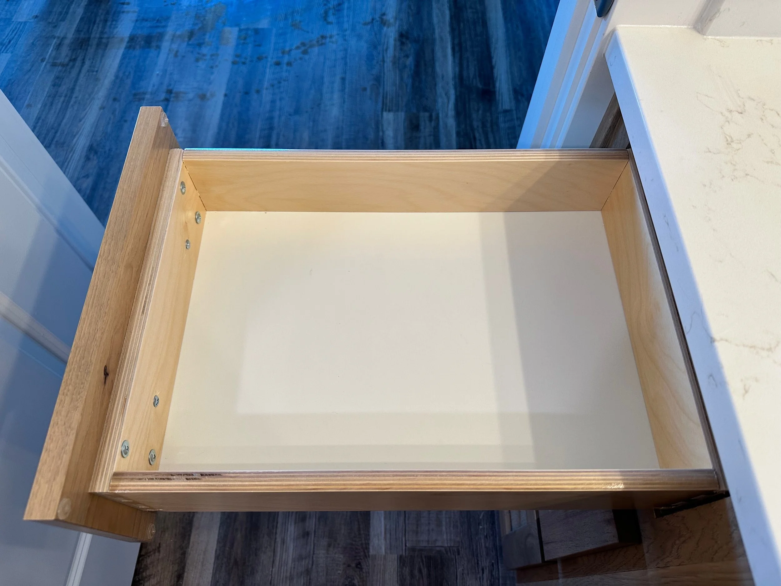 Empty wooden cabinet with a white interior, open with no items inside, situated on a wood floor with a kitchen counter nearby.