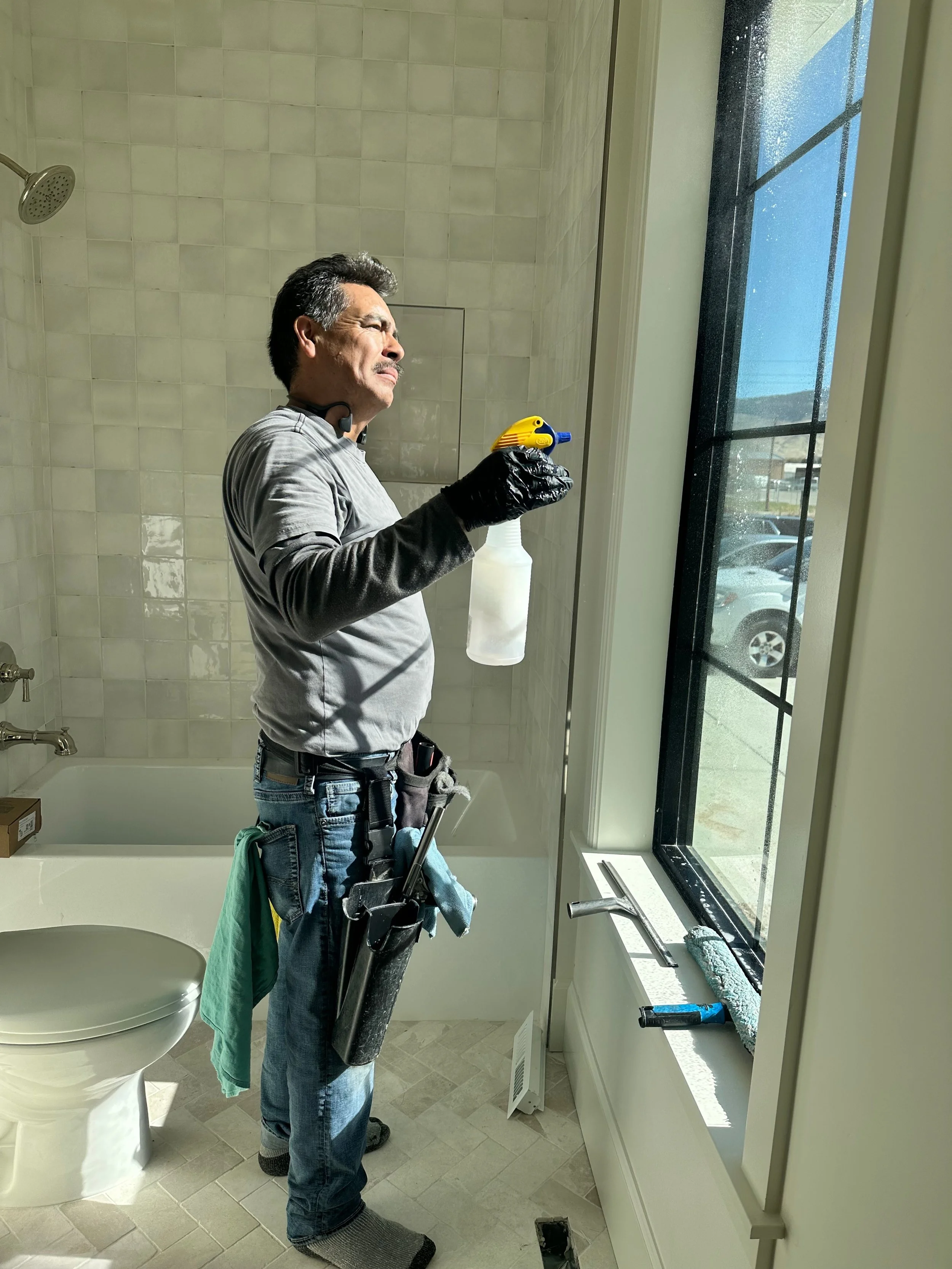 A man standing in a bathroom, wearing gloves and a tool belt, holding a spray bottle and a yellow and blue device, cleaning the window.