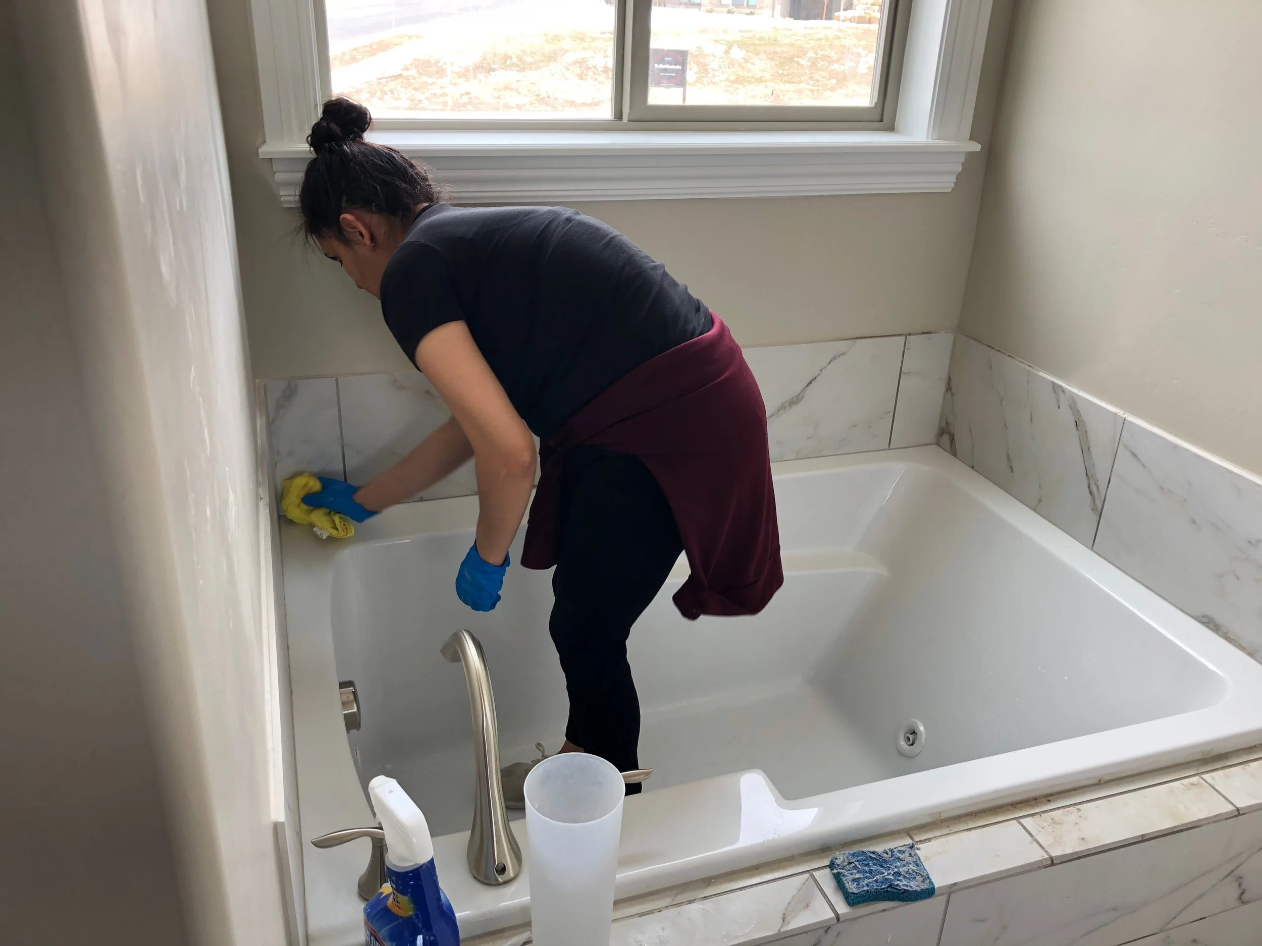Woman cleaning a bathtub with a yellow cloth, wearing blue gloves, inside a bathroom with a large window.