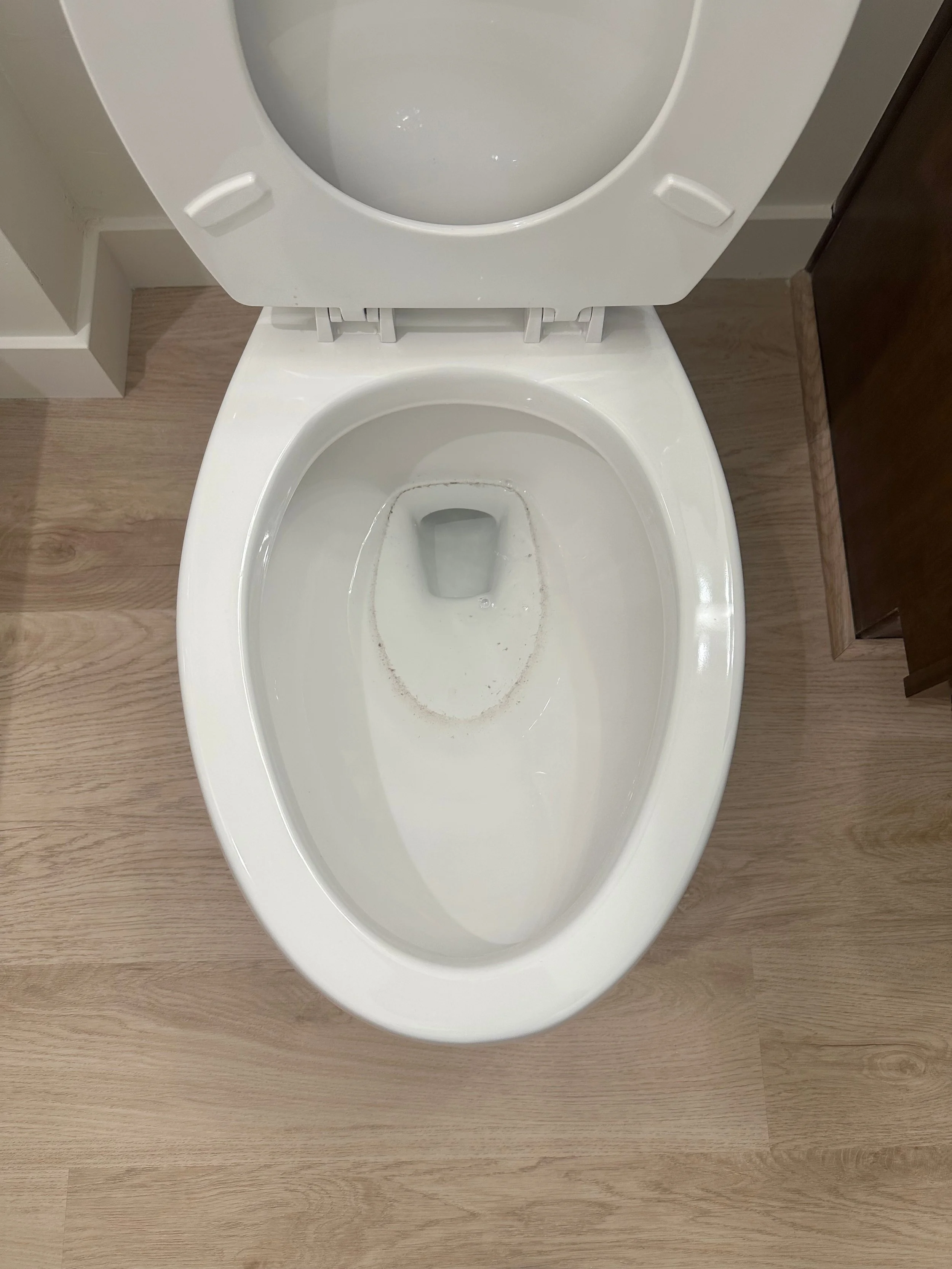 A white toilet with the lid open in a bathroom, showing the inside of the bowl, next to wooden flooring and a cabinet.