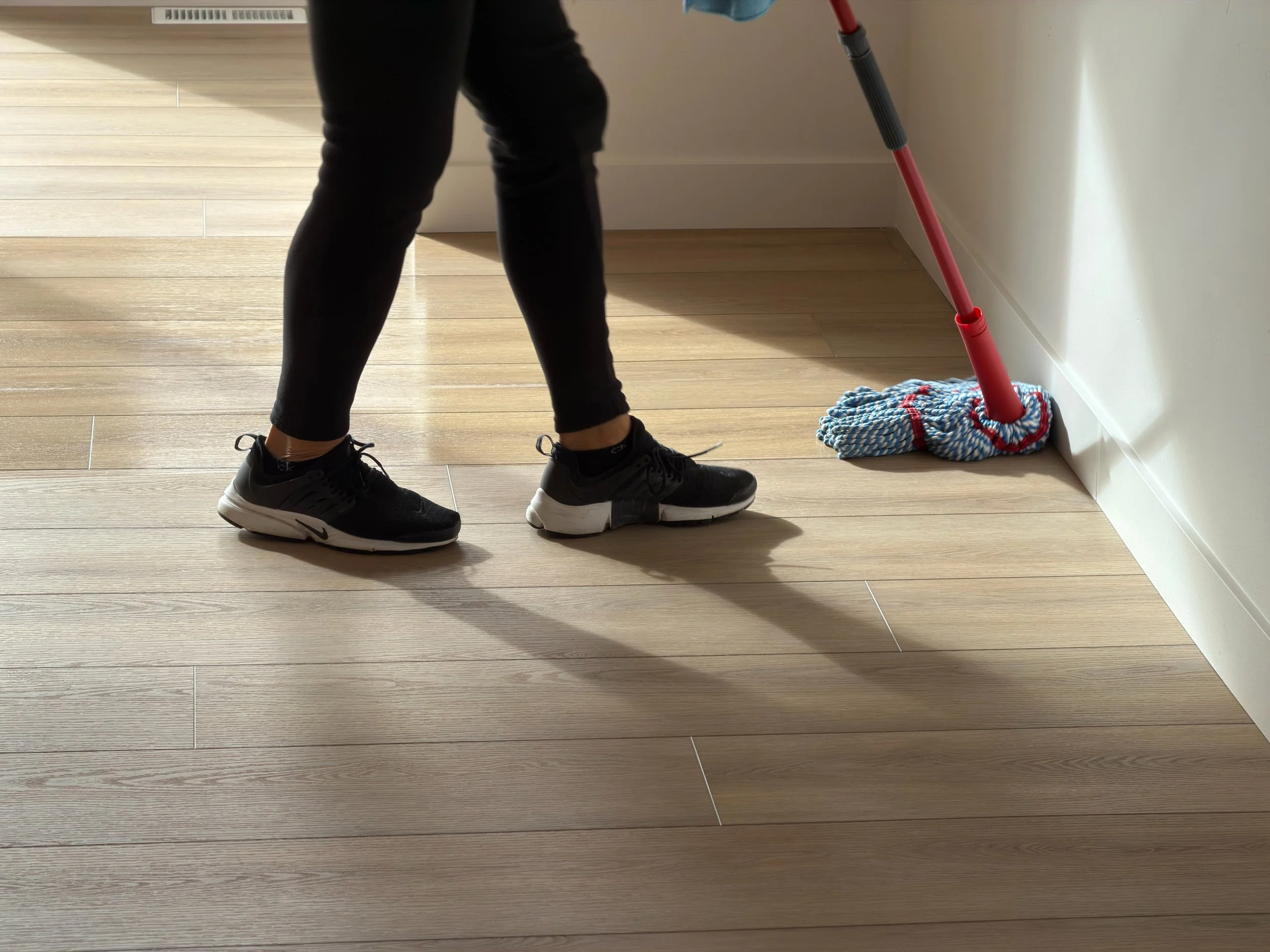 Person in black athletic shoes and black pants mops wooden floor with a red mop and blue and white striped cloth near the wall.