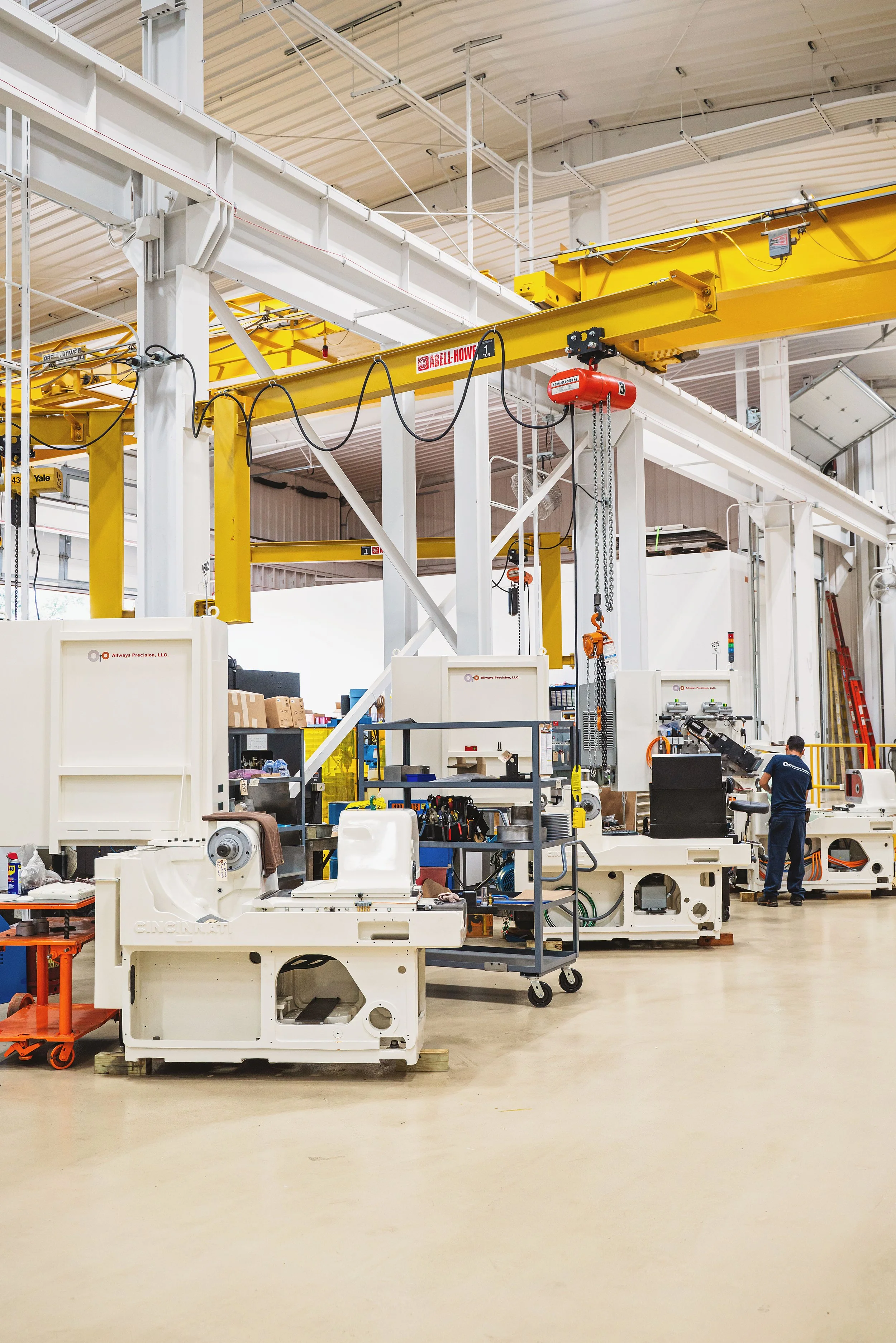 Industrial workshop with machinery, tools, and equipment, including a large yellow overhead crane and a person working on a piece of industrial equipment.