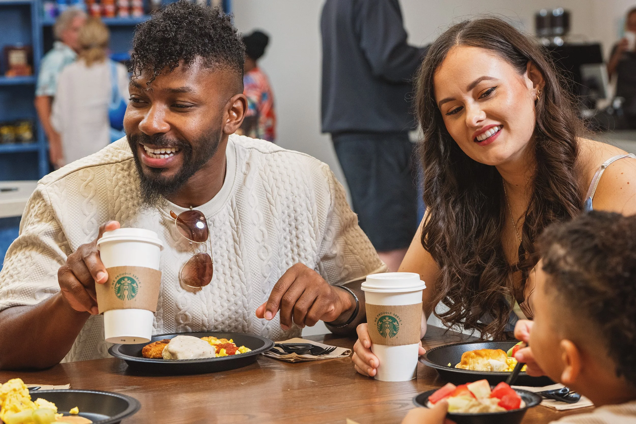 A group of four people sitting at a table with food and drinks, enjoying a meal at Starbucks. Two adults and two children are visible, smiling and engaging with each other.