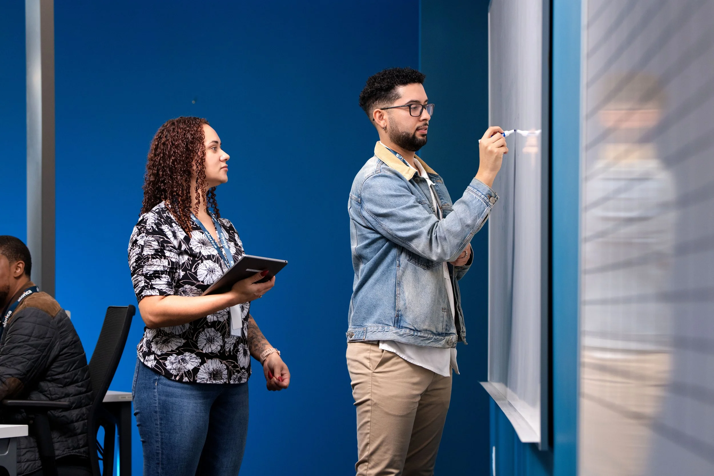 A man and a woman in a classroom or office, with the man writing on a whiteboard and the woman observing while holding a tablet.